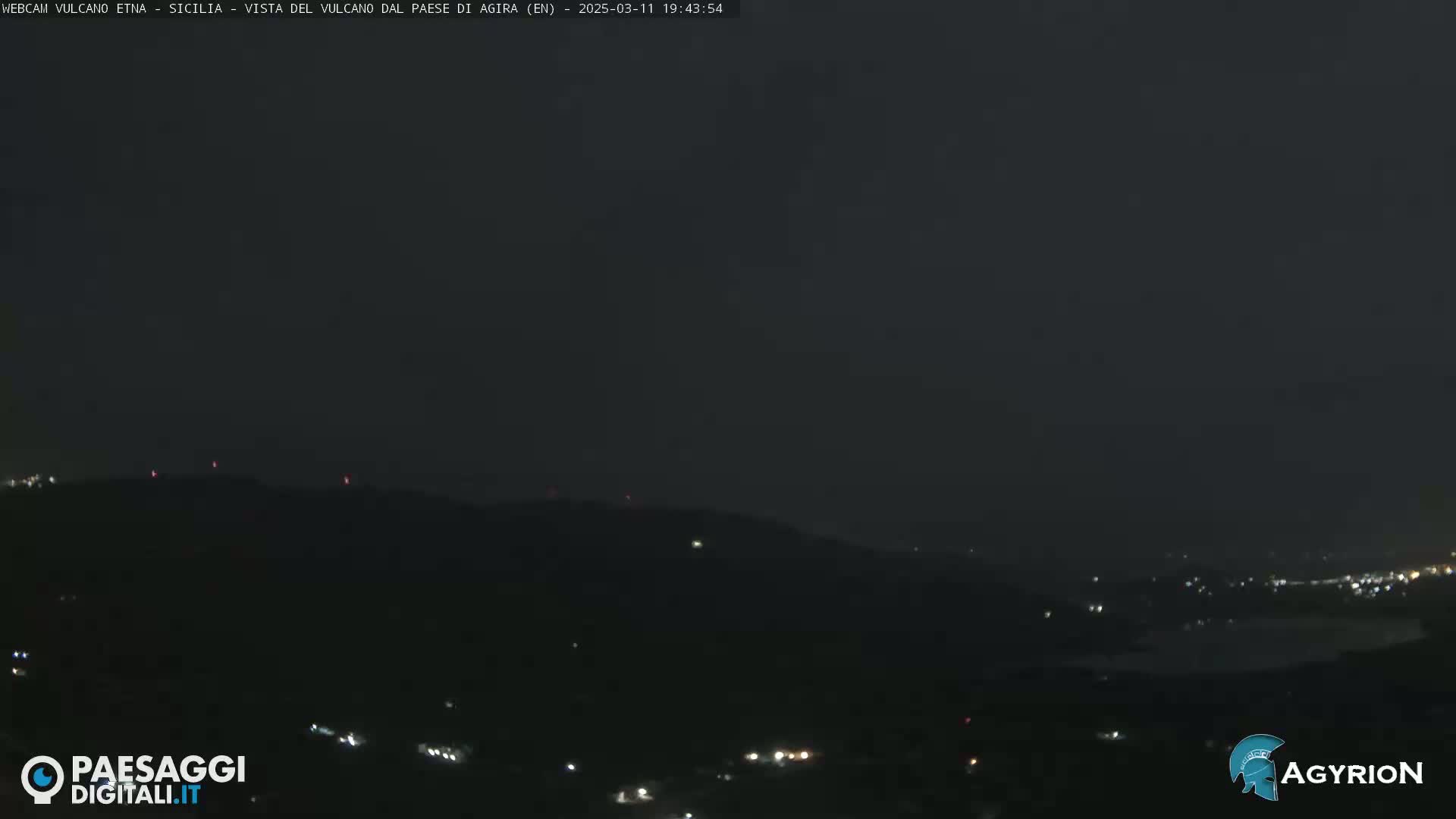 A nighttime long shot of dimly lit hills and a town under a dark sky.