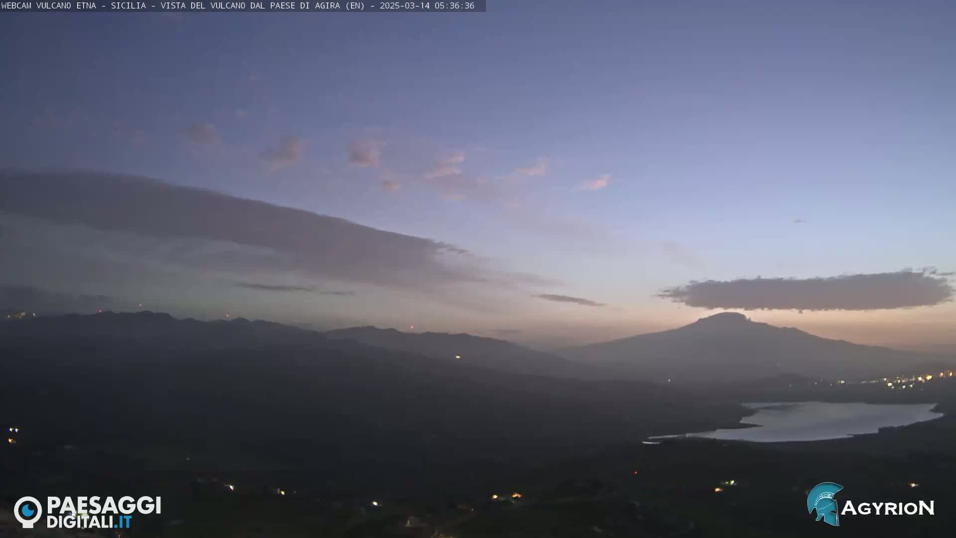 A hazy sunrise illuminates a distant mountain range, a lake, and a valley speckled with faint lights under a twilight sky with thin clouds.