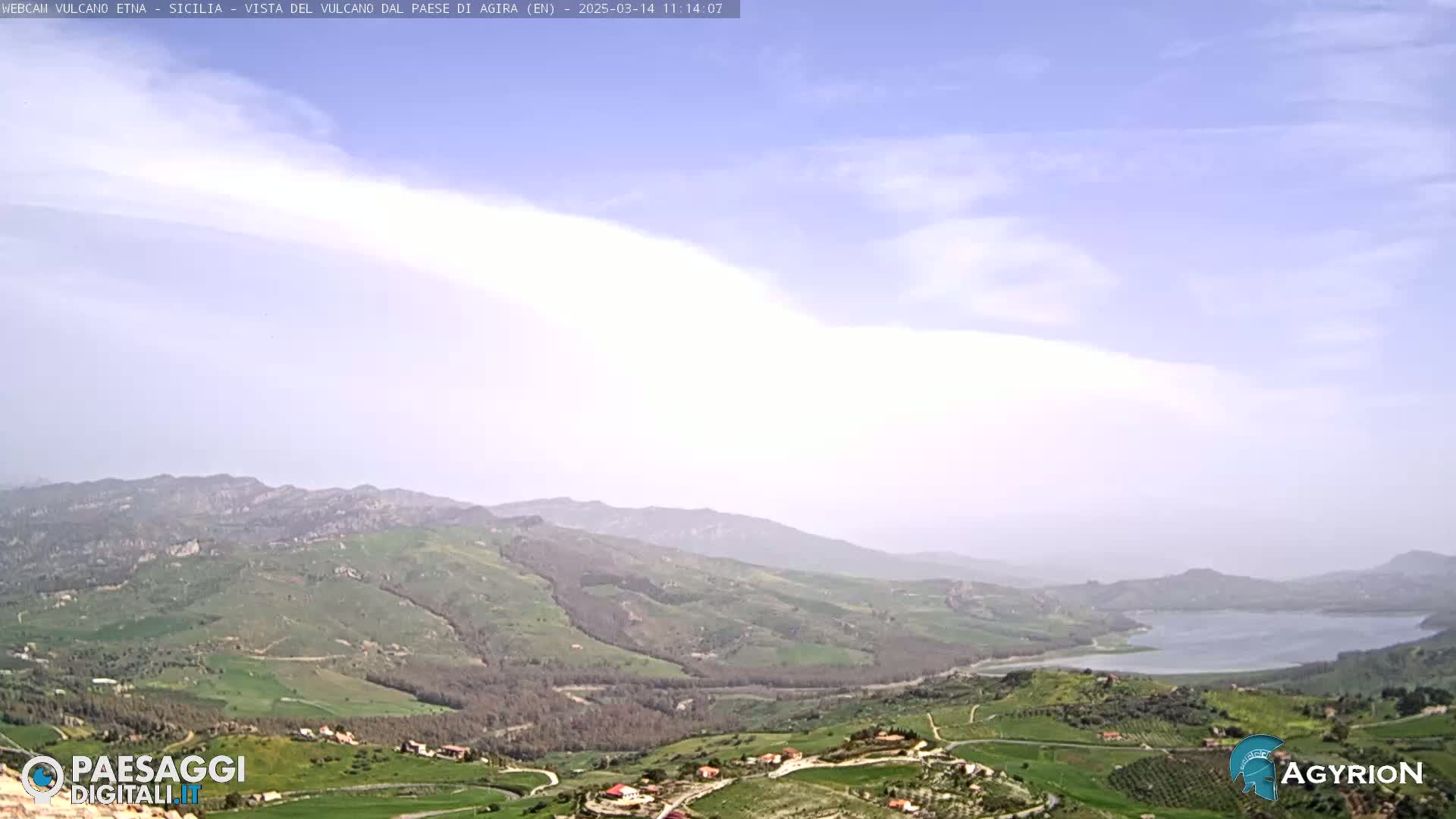 A hazy, partly cloudy day reveals a picturesque valley with rolling green hills, farmland, a lake, and distant mountains.