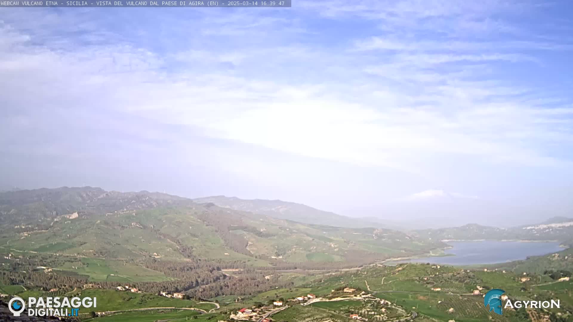 A vast, hilly landscape under a mostly clear sky with some thin clouds stretches to a lake in the distance.