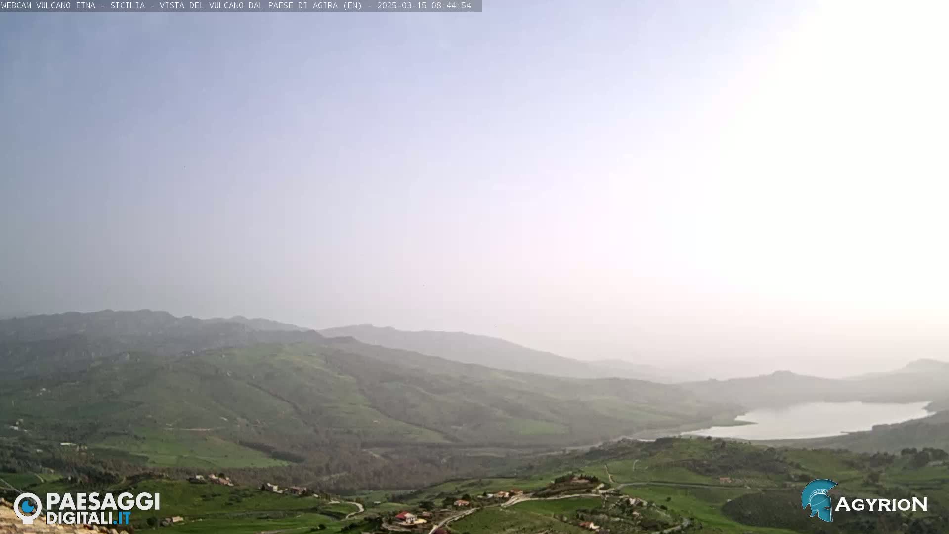 A hazy, light-colored sky overlooks a vast, green valley with a lake and small villages nestled amongst rolling hills.