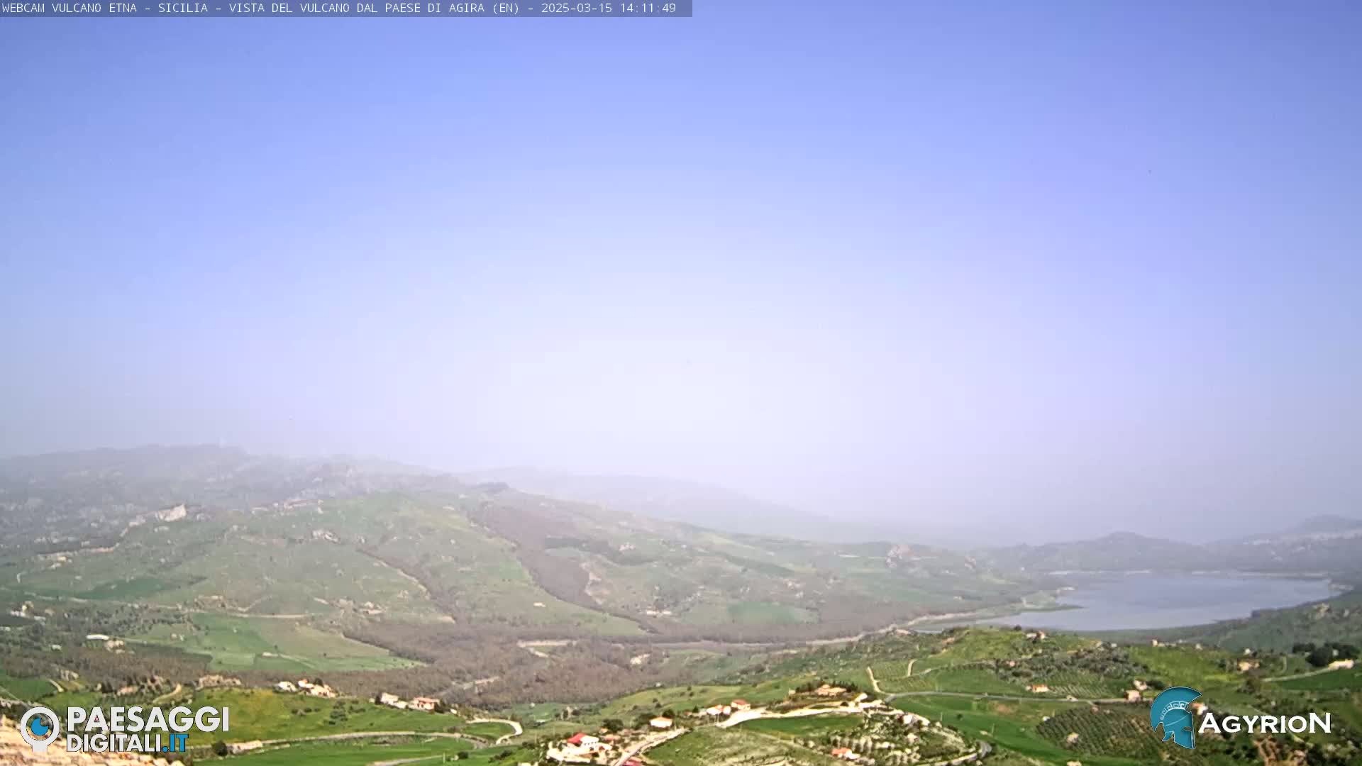 A hazy, long-distance view of a valley with rolling green hills, scattered buildings, and a lake under a clear, light-blue sky.