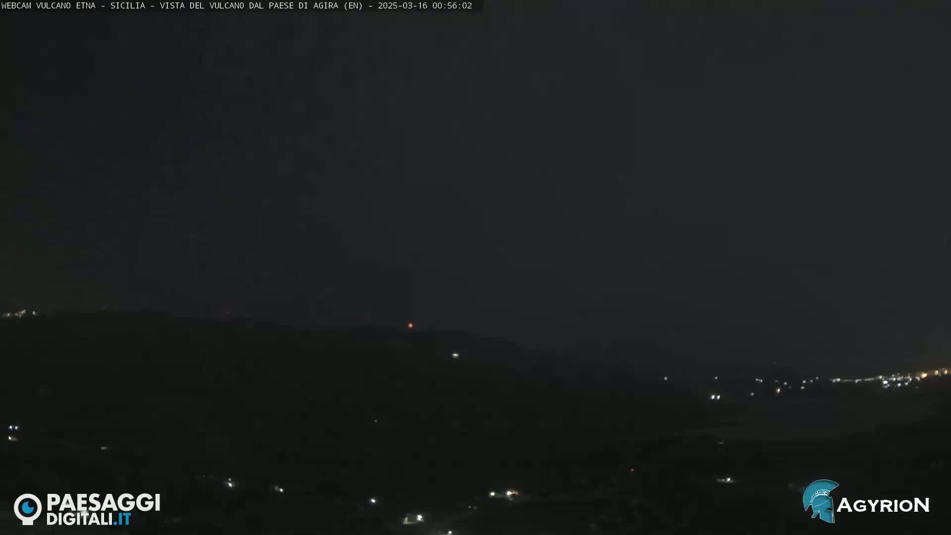 A nighttime long shot of a dark, mountainous landscape with a few scattered lights visible in the distance.