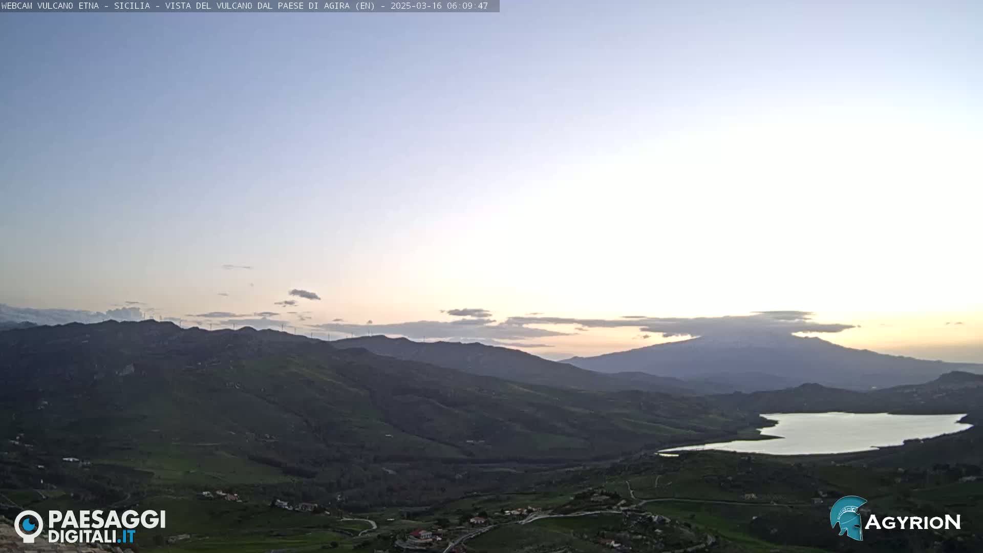 A tranquil, sunlit view of rolling hills, a lake, and a distant mountain range under a pale, clear sky.