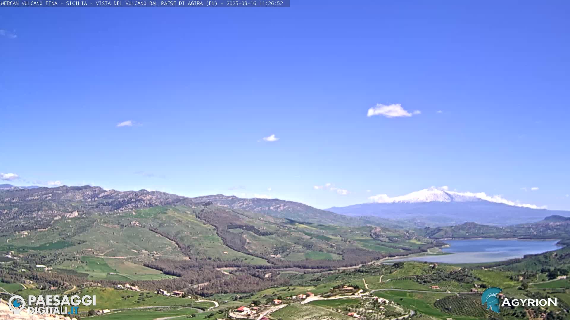 A mostly sunny day reveals a vast, verdant valley with a lake, rolling hills, and a snow-capped mountain in the distance.