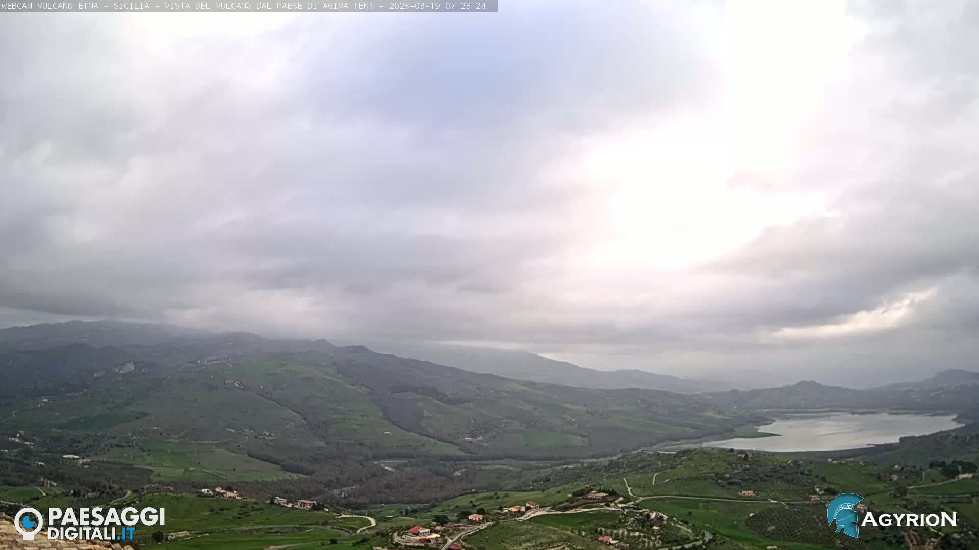 A vast, hilly landscape under an overcast sky features a lake nestled within the rolling green hills and farmland.