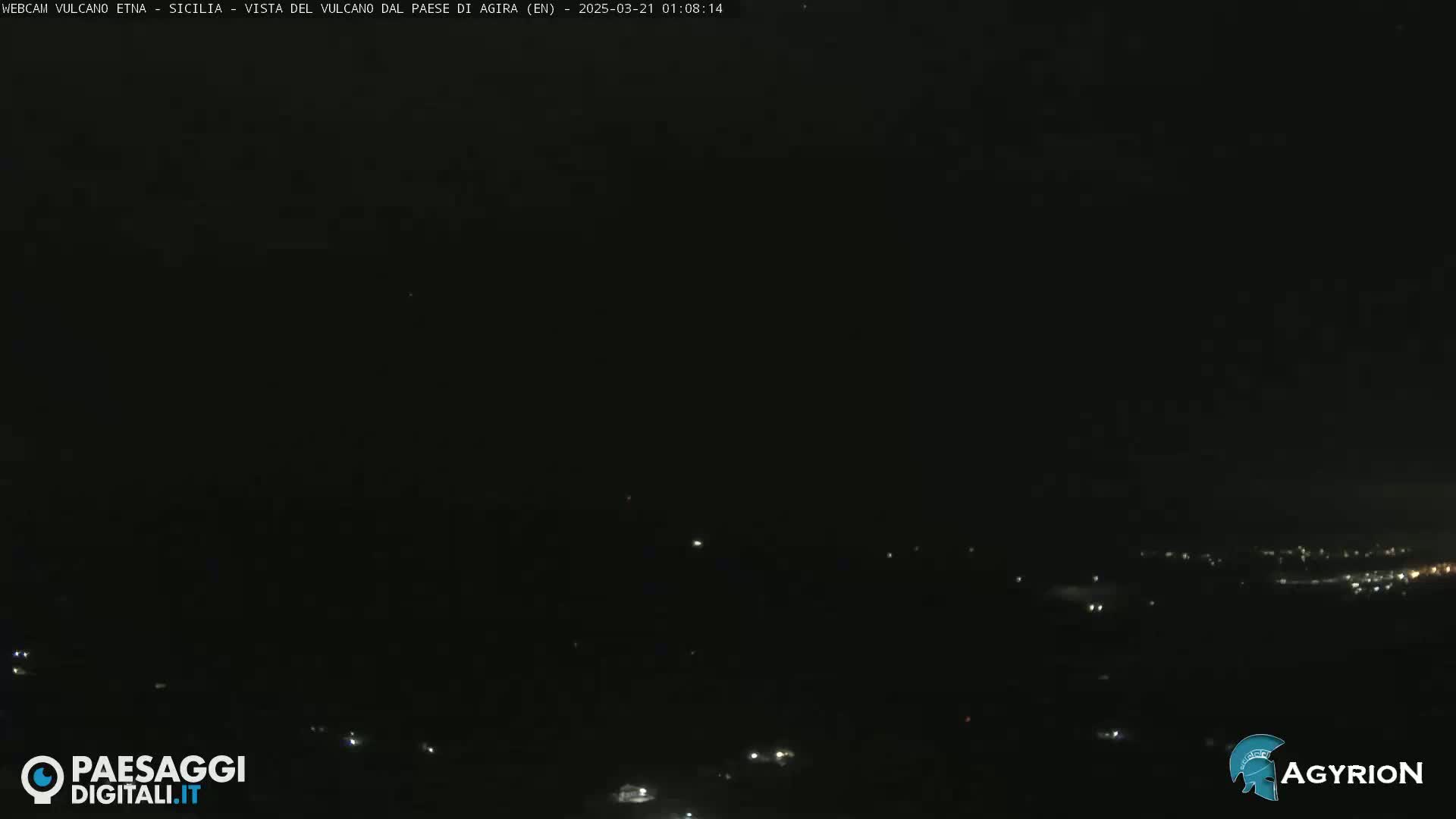 A nighttime, long shot of a dark landscape with a few scattered lights visible in the distance.
