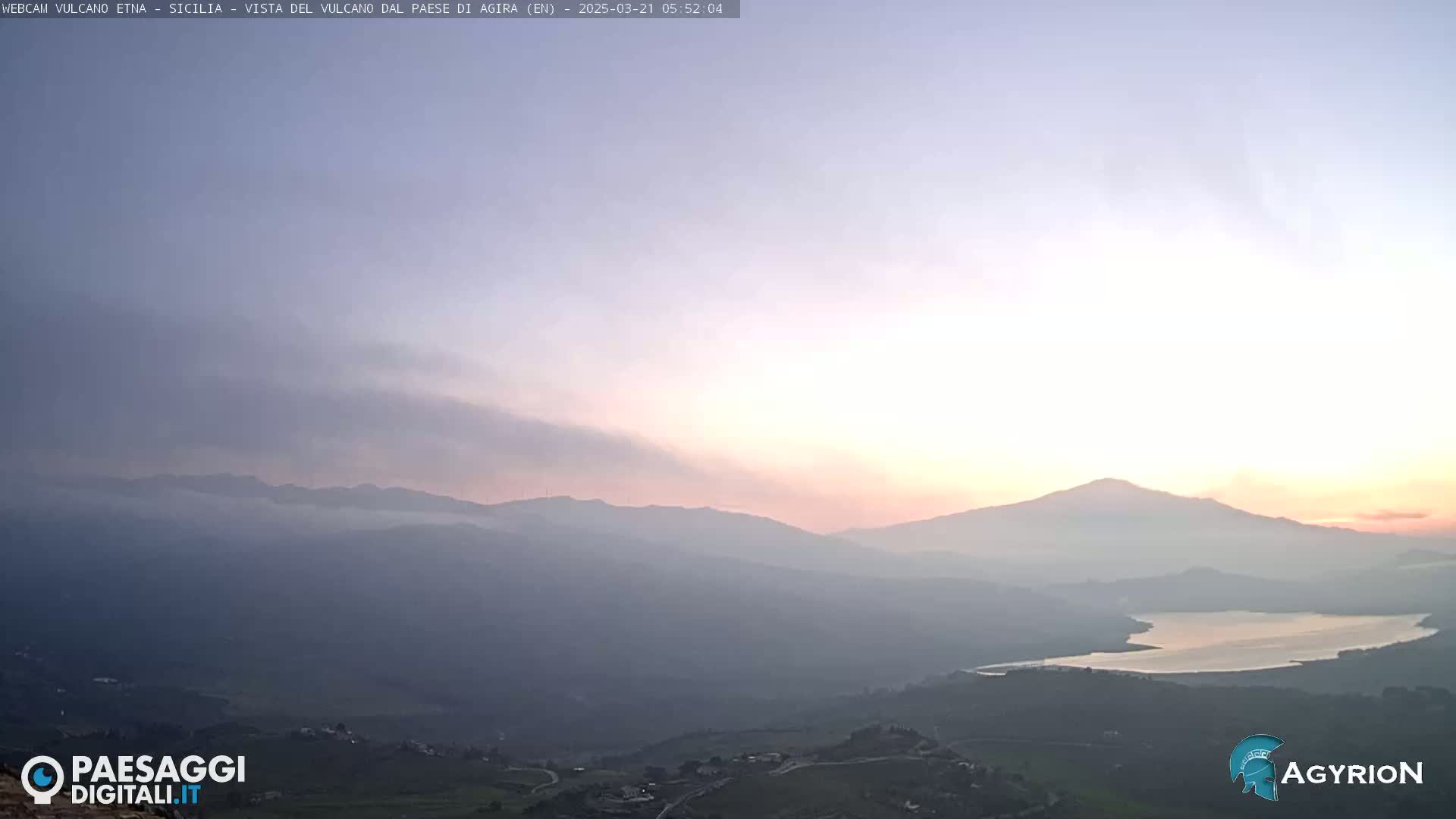 A hazy sunrise illuminates a mountain range and a lake in the distance, with a valley and scattered buildings in the foreground.