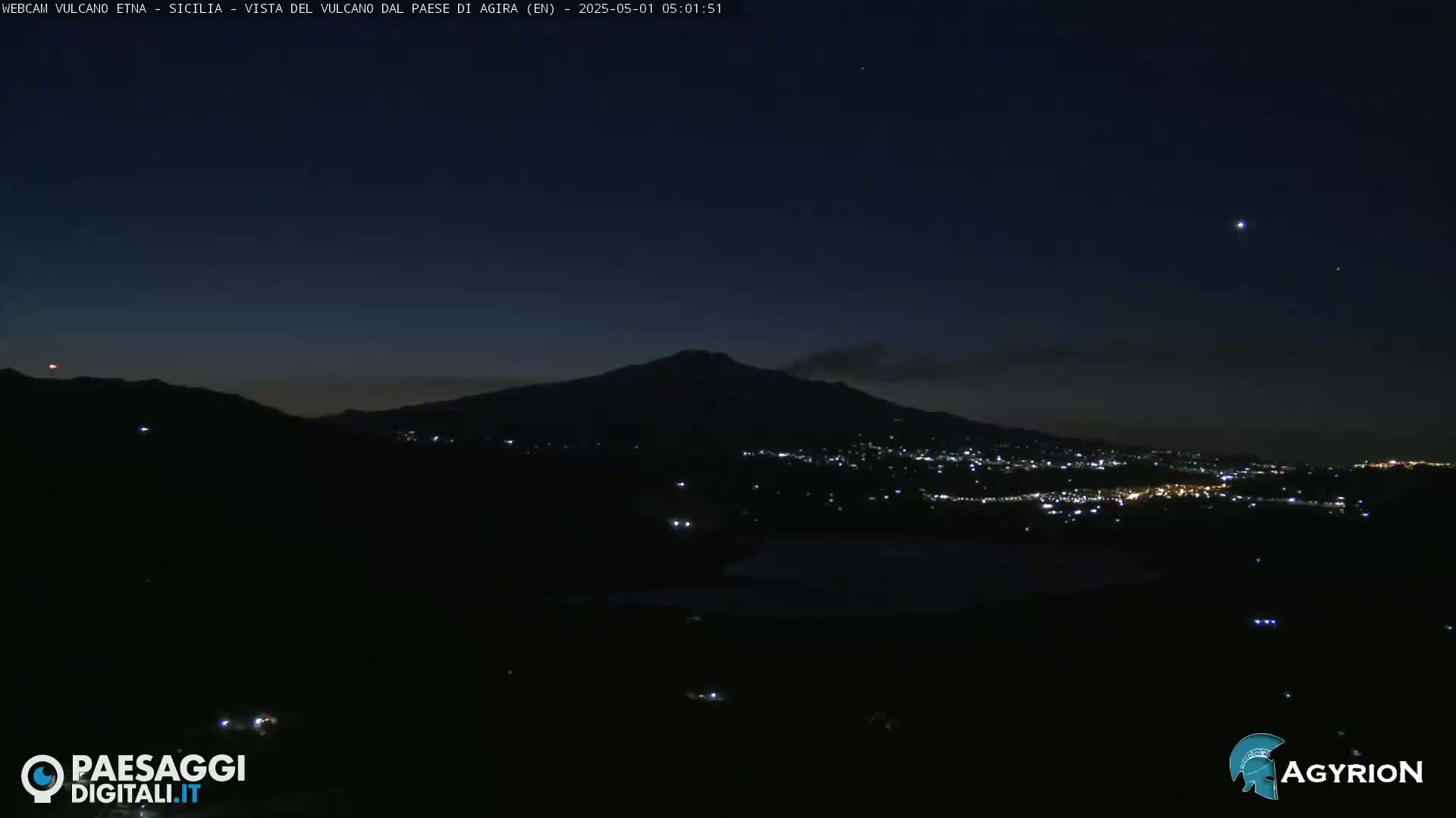 A dark, nighttime view of a city illuminated by lights at the base of a large, silhouetted mountain under a mostly clear, starlit sky.
