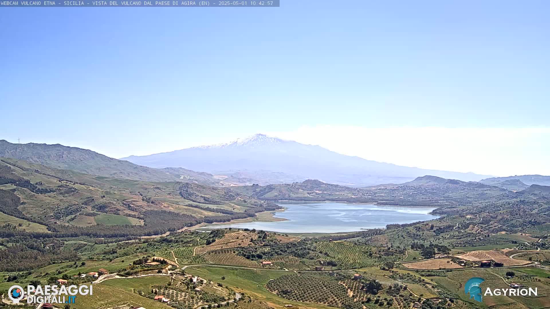 A snow-capped mountain is visible in the distance, overlooking a valley of green hills, farmland, and a lake under a clear blue sky.