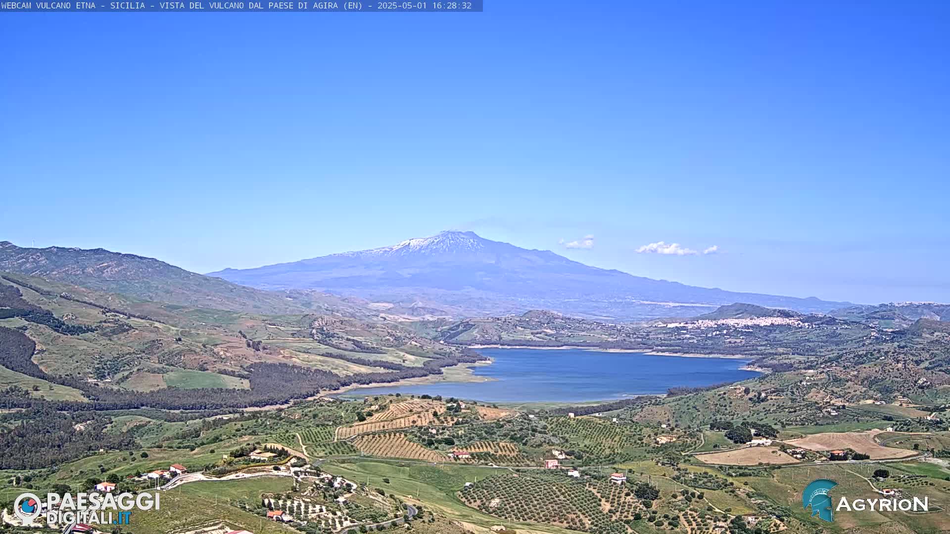 A snow-capped volcano is visible in the distance over a hilly landscape with a lake and cultivated fields under a clear blue sky.