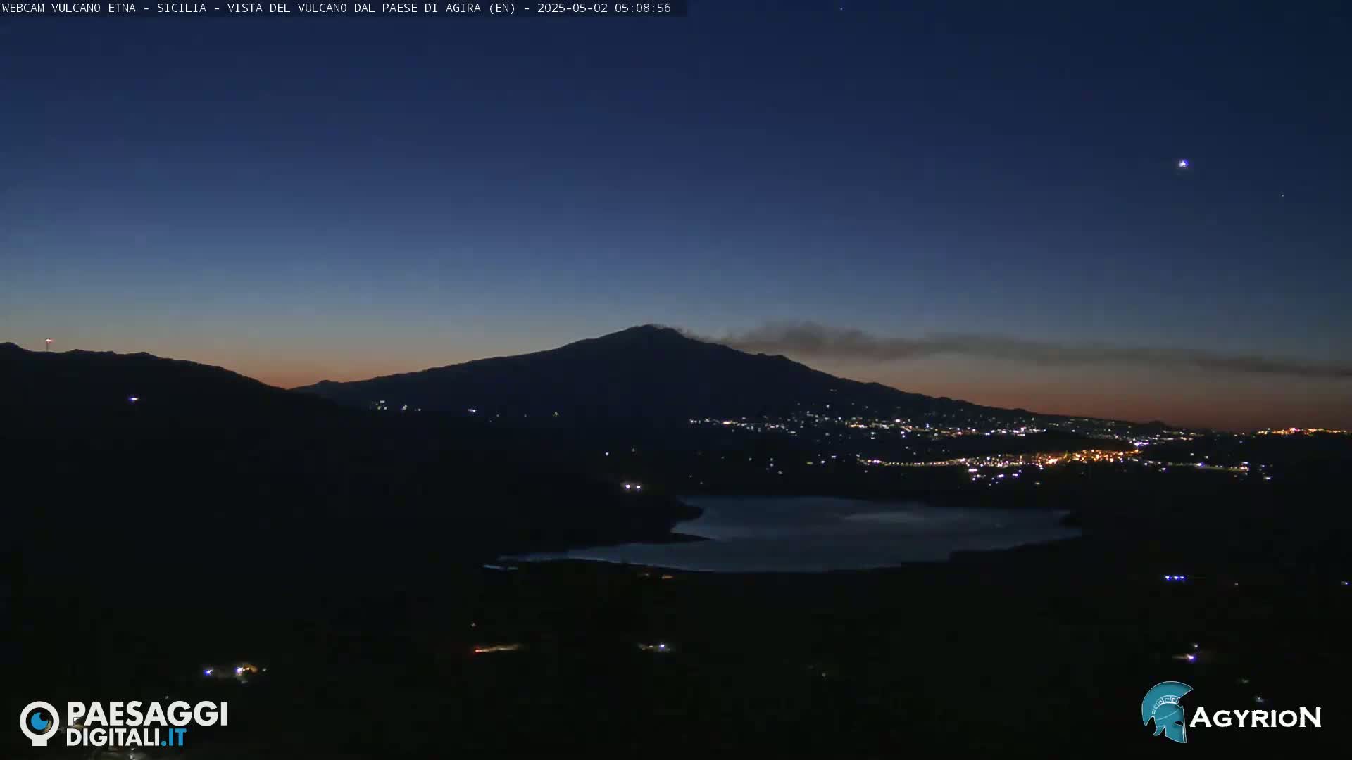 A dark, nighttime landscape shows a volcano emitting smoke under a twilight sky, with a town's lights visible below and a body of water in the foreground.