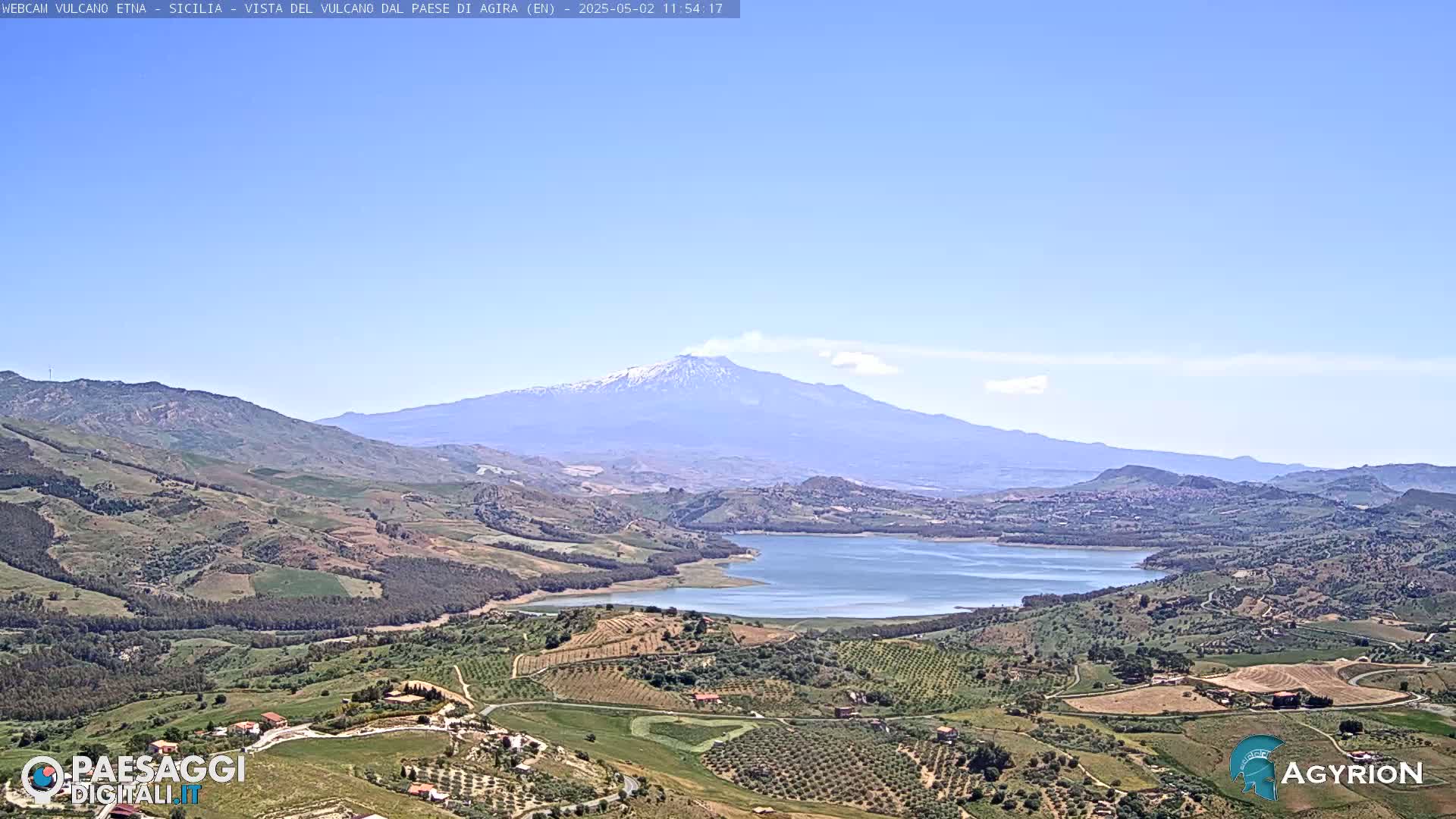 A snow-capped mountain is visible in the distance, overlooking a valley with a lake and farmland under a clear blue sky.