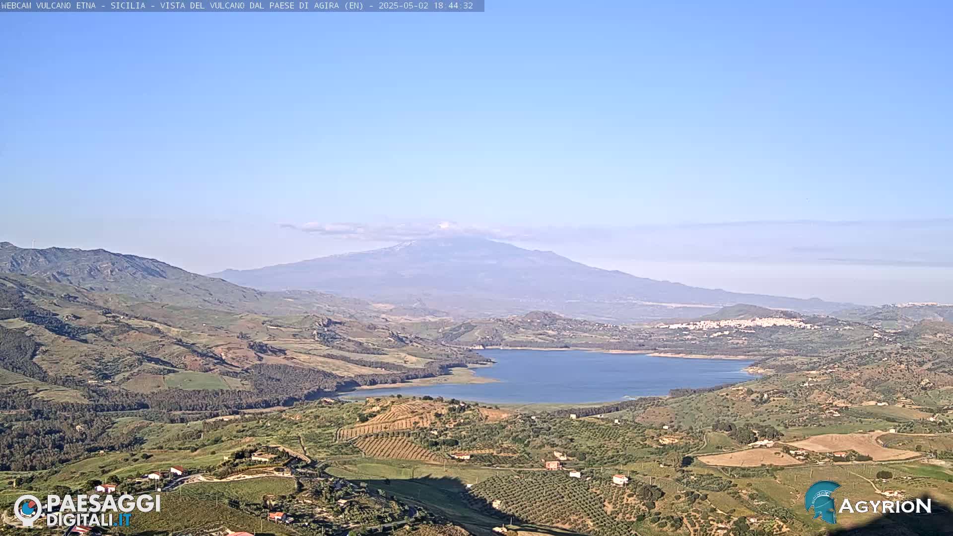 A distant, hazy mountain is visible beyond a valley of cultivated fields and a lake under a clear sky.