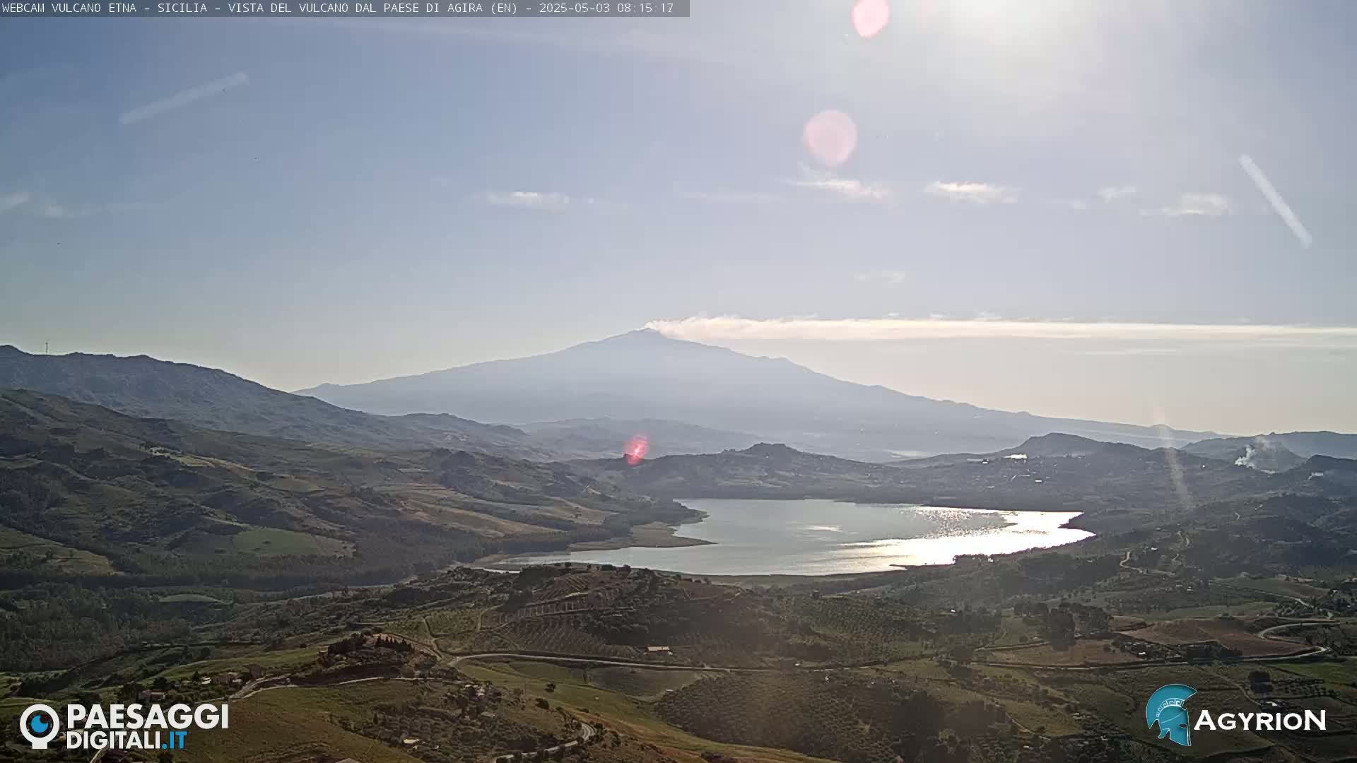 A sunlit view of a large lake nestled among rolling hills, with a distant, hazy mountain range including a volcano under a mostly clear sky.