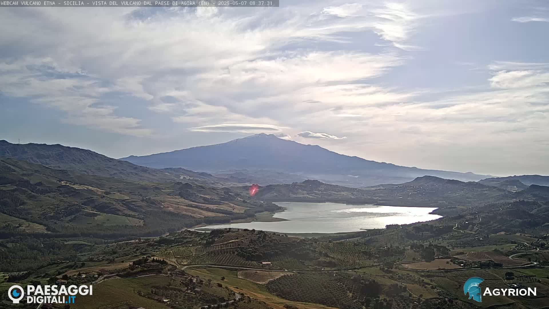 A partly cloudy sky overlooks a valley with a lake and rolling hills, culminating in a large, snow-capped mountain in the distance.