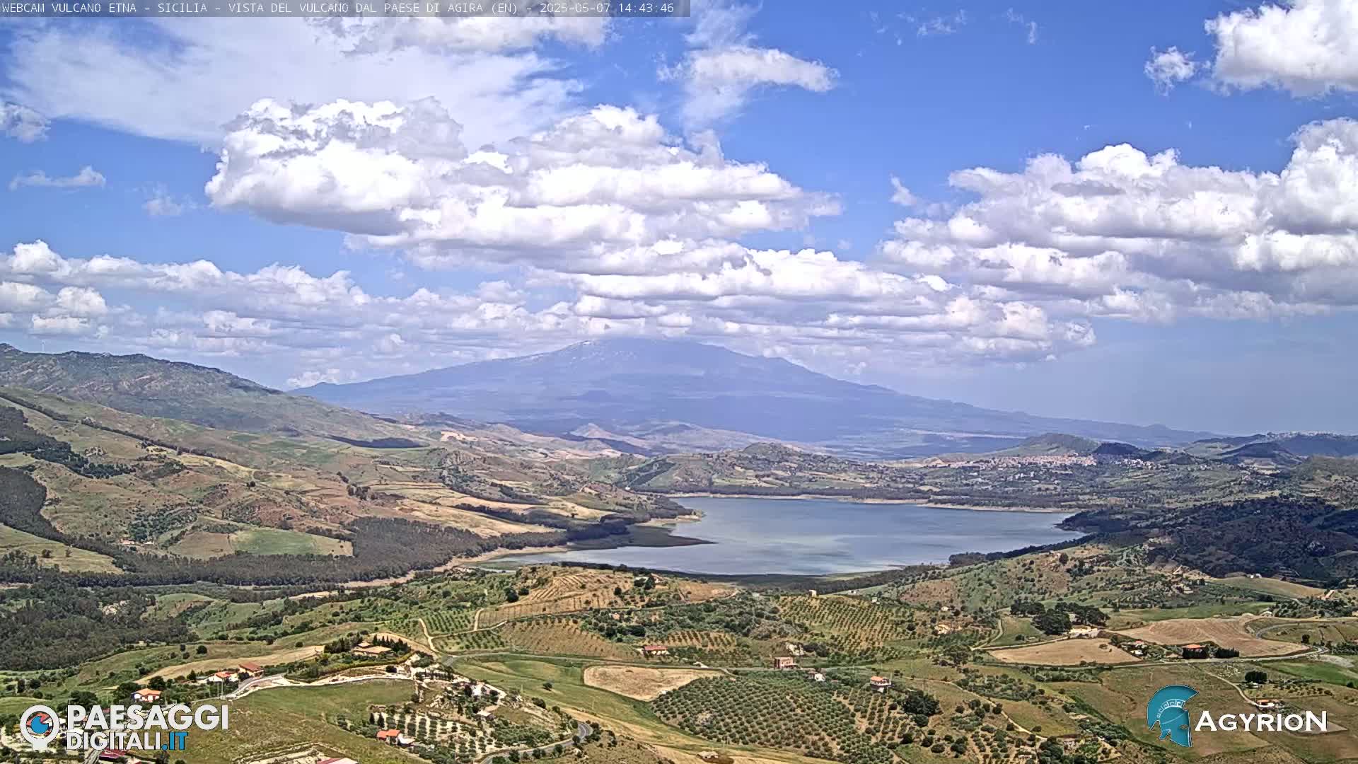 A partly cloudy sky overlooks a vast, hilly landscape with a large lake and a distant, snow-capped mountain.