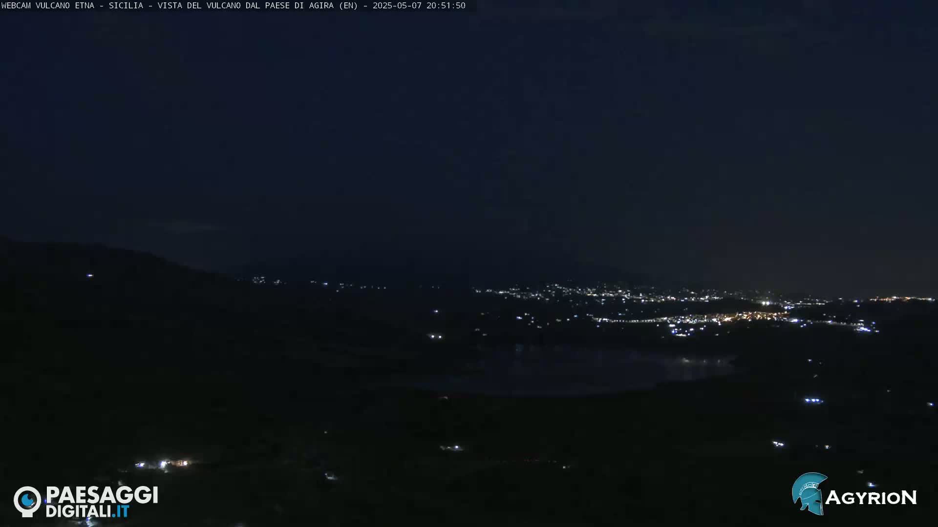 A nighttime, long shot of a city’s lights spread across a dark valley, viewed from a hilltop.