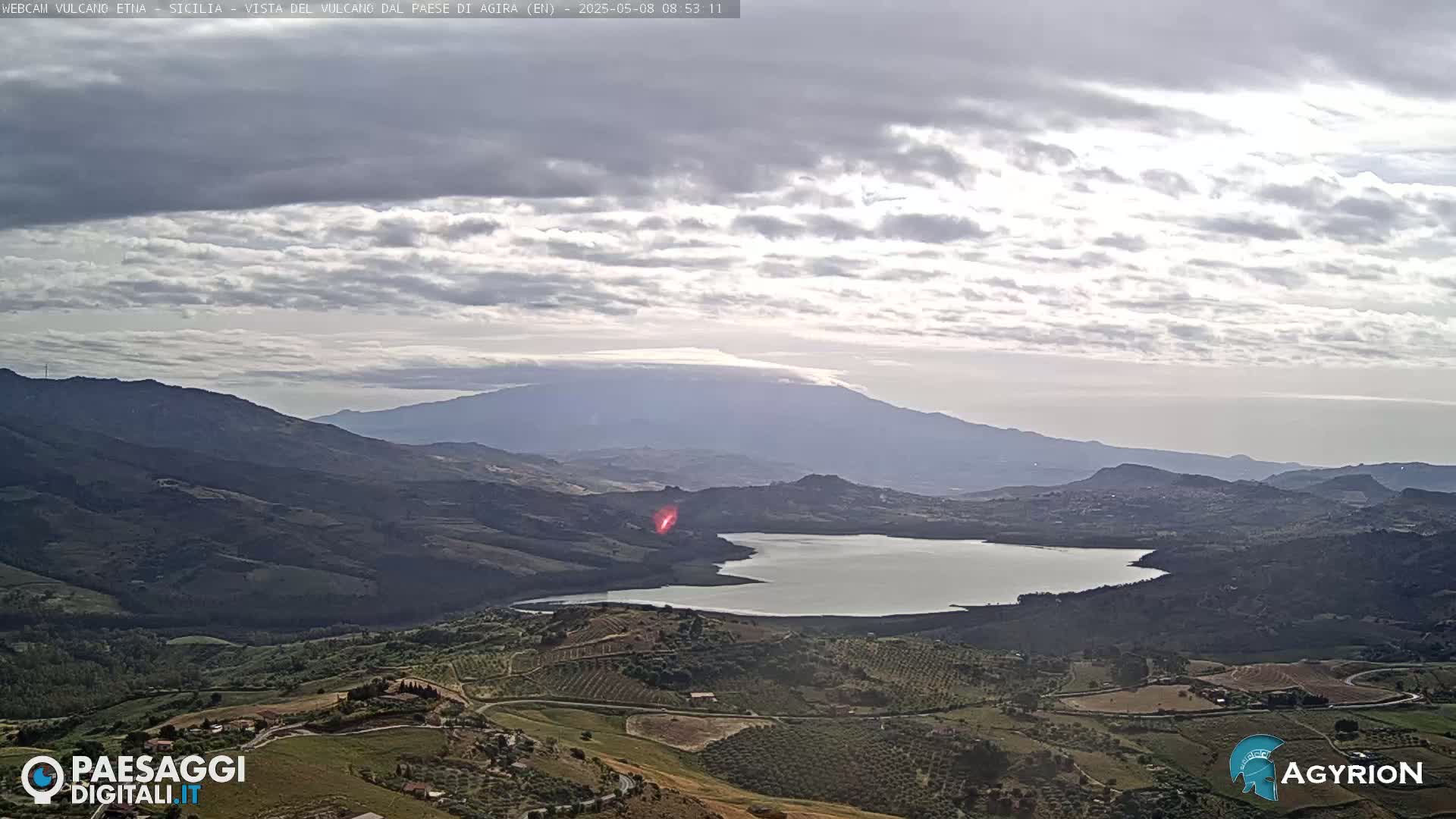 A partly cloudy sky overlooks a vast, hilly landscape containing a lake, with a large mountain in the distance.