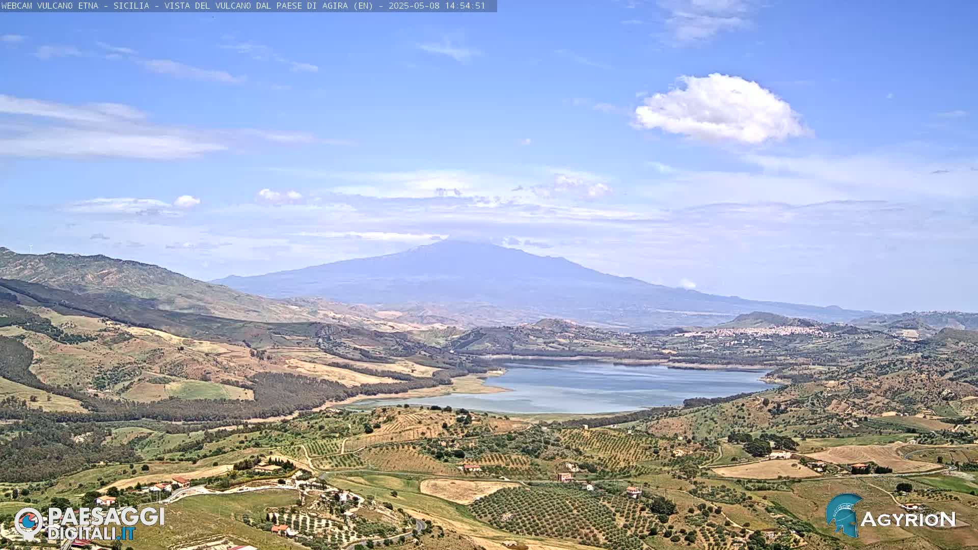 A partly cloudy sky overlooks a vast landscape of rolling hills, farmland, and a lake, with a large, distant mountain in the background.