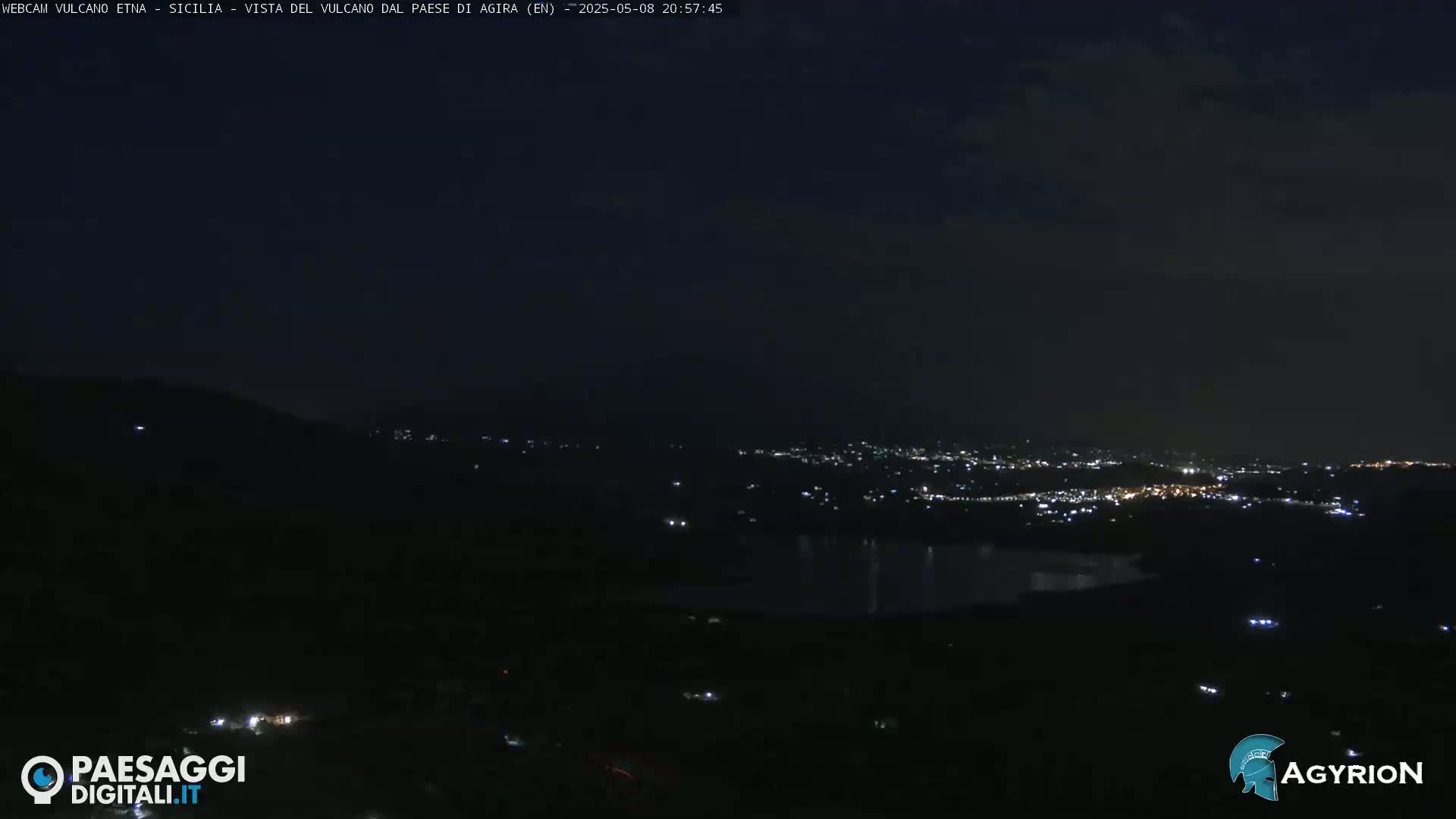 A nighttime, long shot of a city illuminated below a dark sky, with a body of water visible between the city and the vantage point.
