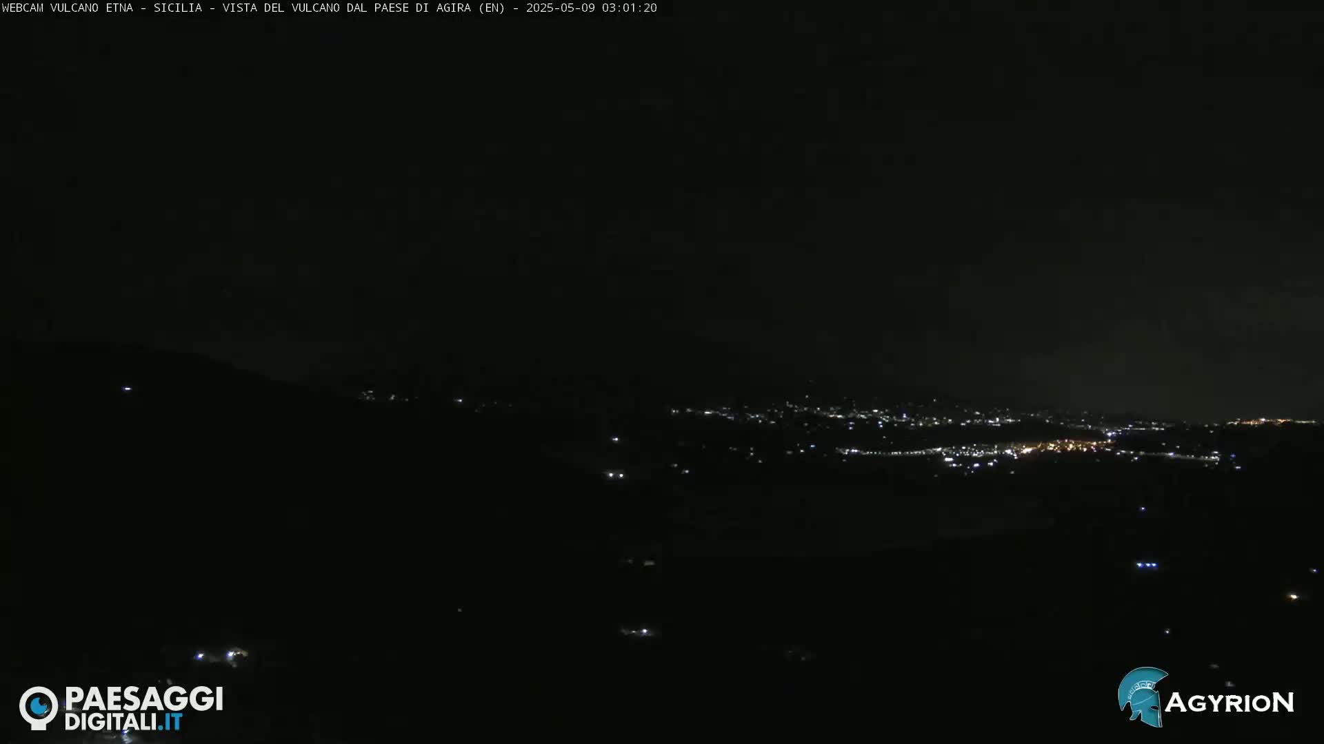 A nighttime long shot shows a dark landscape with a distant city illuminated by scattered lights under a clear, dark sky.