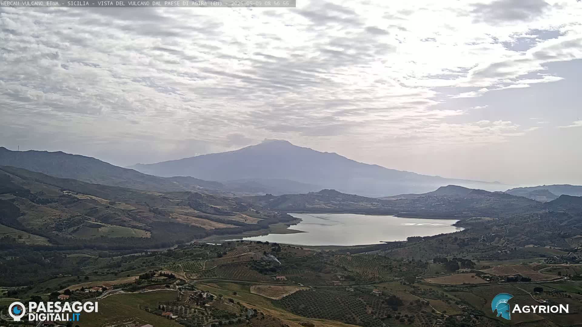 A hazy, partly cloudy day reveals a distant, large mountain range with a lake in the foreground, and rolling hills and farmland stretching across the landscape.