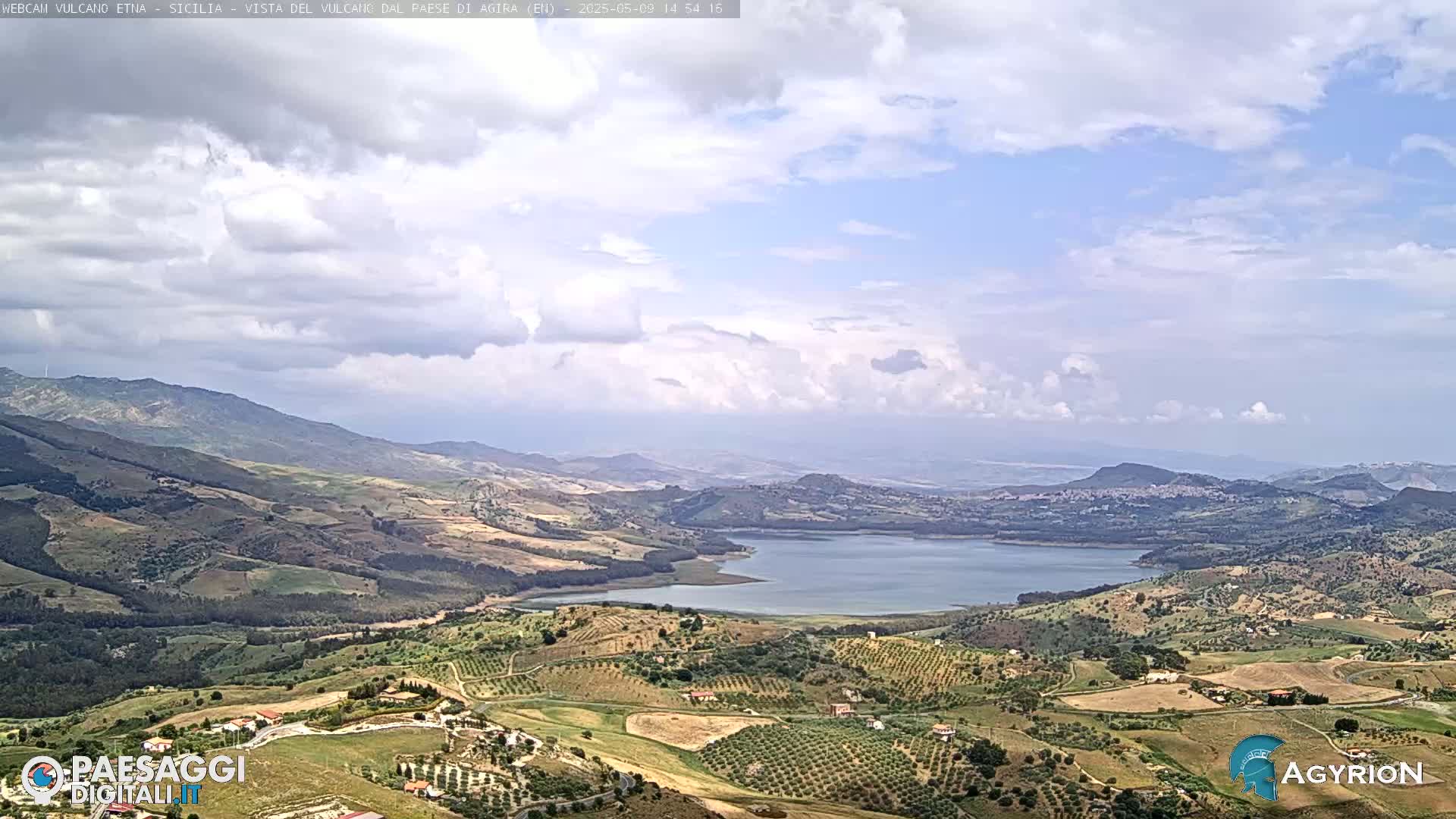 A partly cloudy day reveals a vast, hilly landscape featuring a large lake nestled among cultivated fields and distant mountains.
