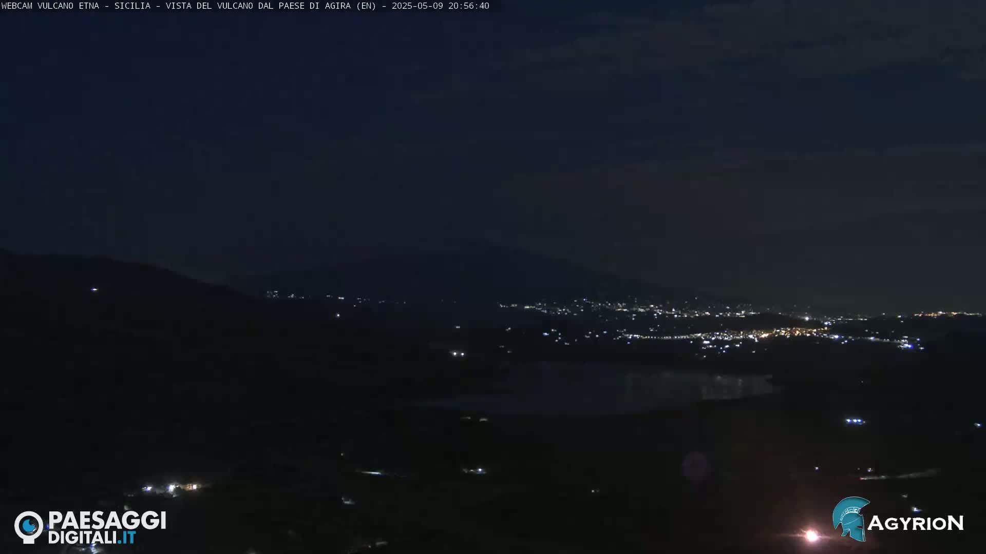 A nighttime, long shot of a city's lights nestled in a valley below a dark mountain range under a mostly clear sky.