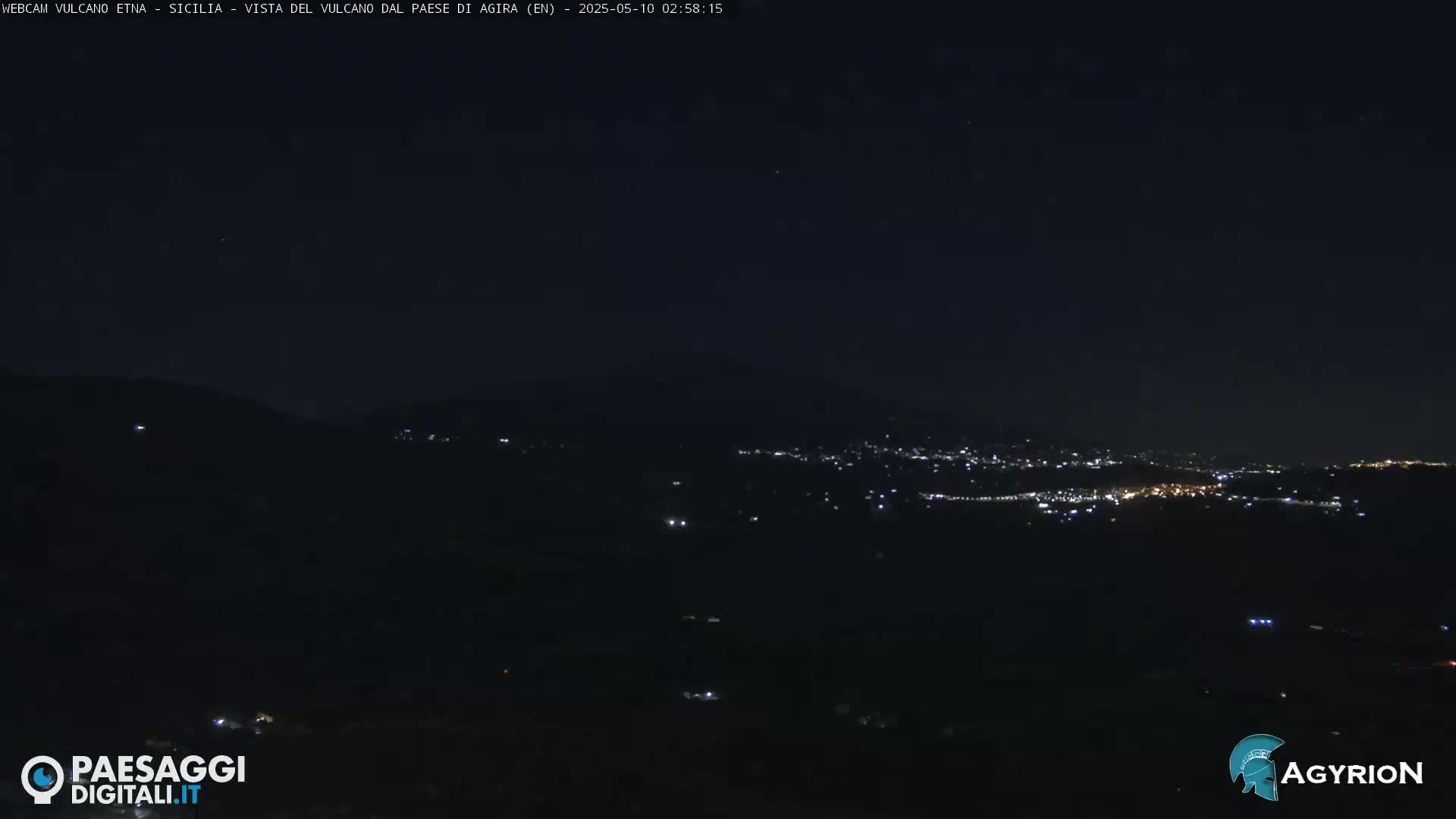 A nighttime long shot shows a dark, hilly landscape with a distant town illuminated by scattered lights under a clear, dark sky.