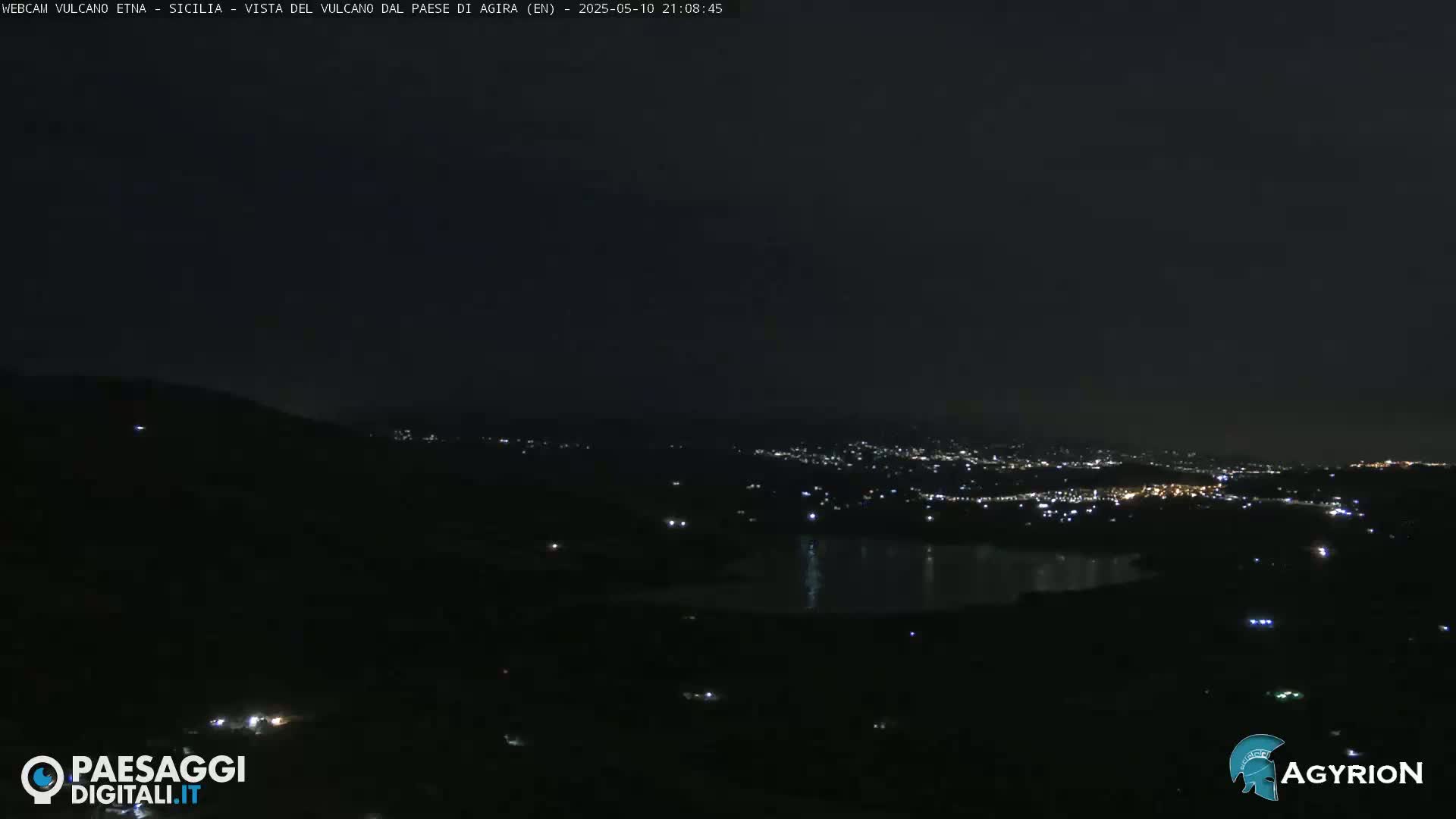 A nighttime, long shot shows a dark, hilly landscape with a body of water reflecting city lights in the distance.