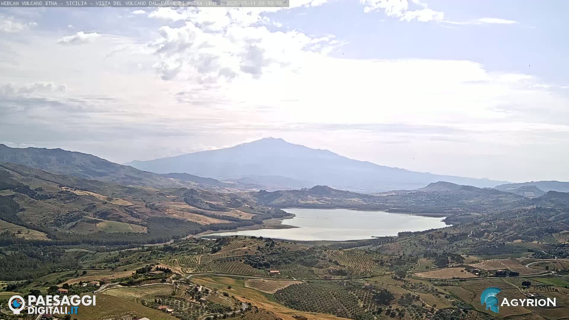 A partly cloudy day reveals a vast, hilly landscape with a large lake and a distant, hazy mountain range.