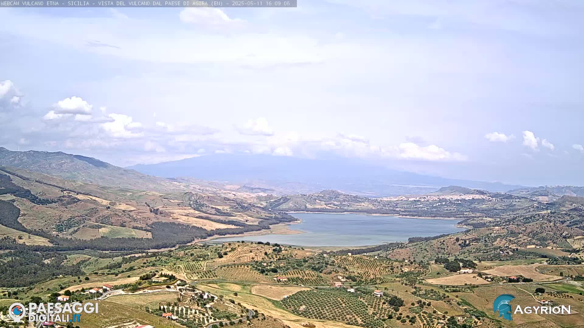 A partly cloudy day reveals a vast, hilly landscape with a lake nestled among rolling green and brown fields, mountains in the background.