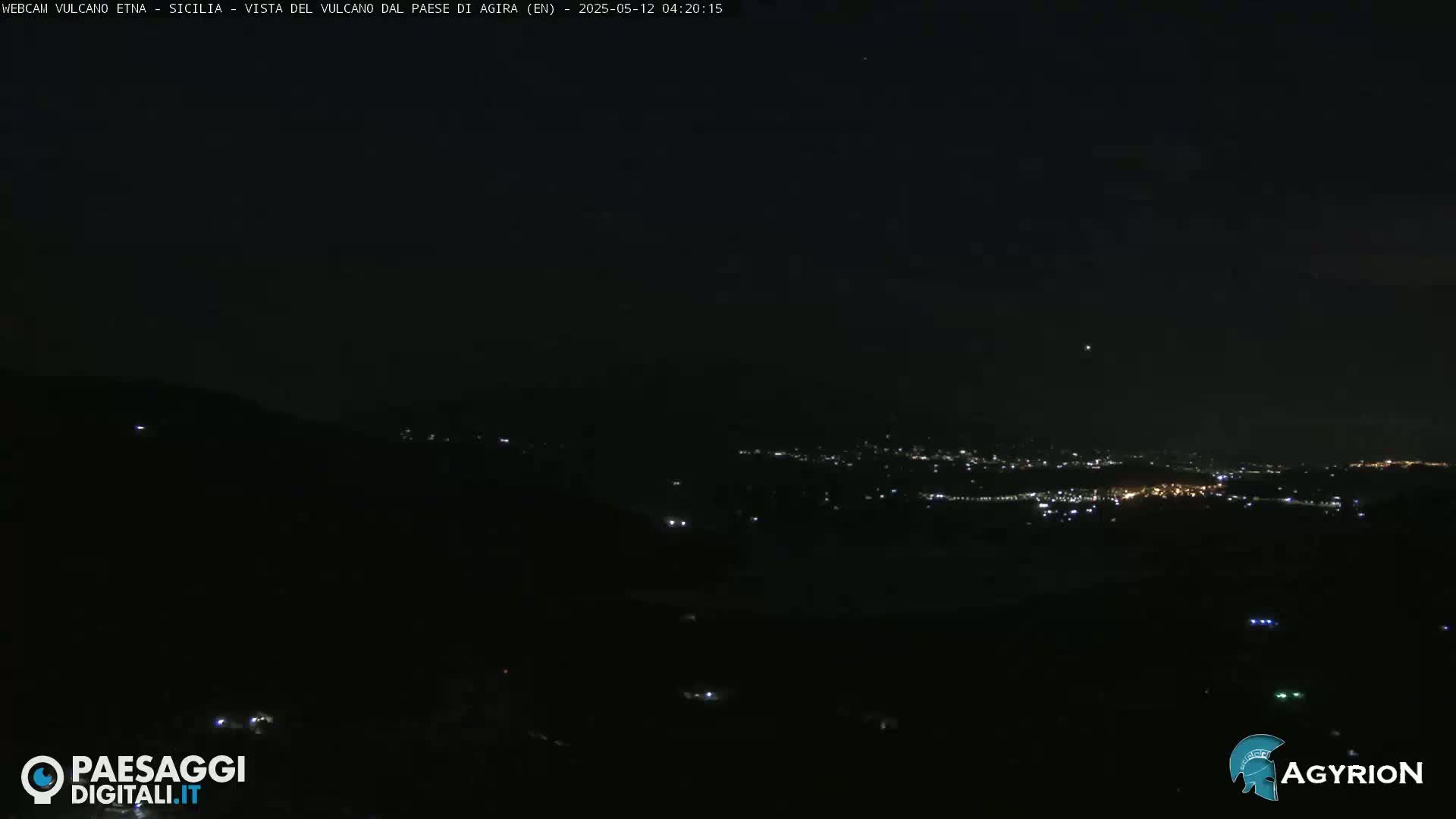 A nighttime long shot shows a dark landscape with a distant town illuminated by lights under a clear, dark sky.