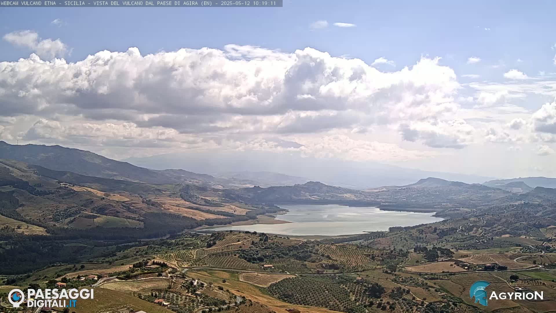 A partly cloudy sky overlooks a vast, hilly landscape containing a lake and cultivated fields.