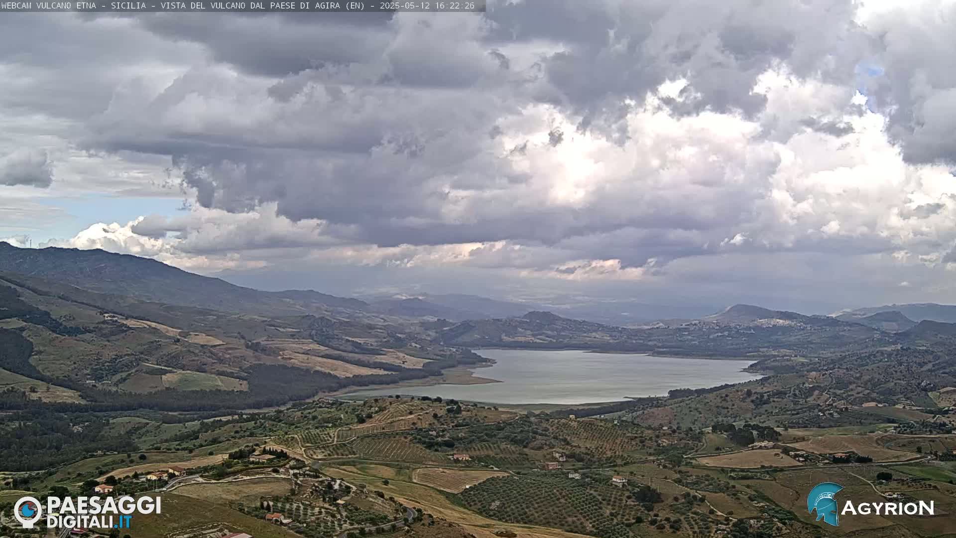 A vast, hilly landscape under a cloudy sky features a lake nestled among rolling green hills and farmland.