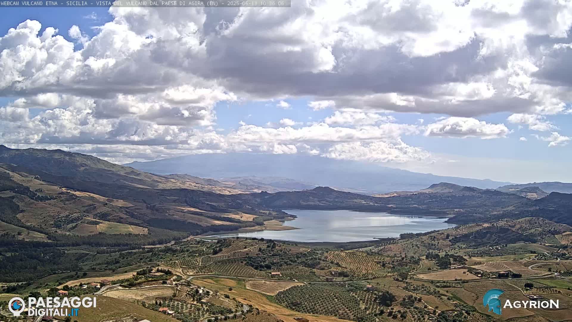 A partly cloudy sky overlooks a lake nestled in a valley between rolling hills and mountains.