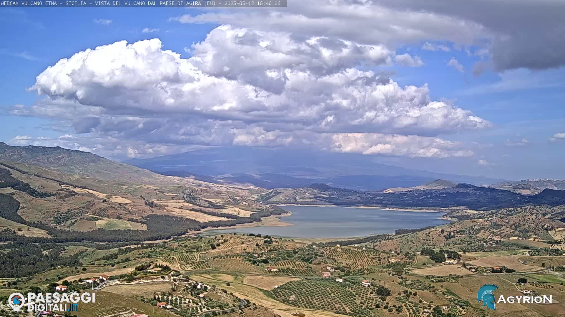A partly cloudy sky overlooks a vast, hilly landscape with a large lake nestled amongst rolling fields and distant mountains.