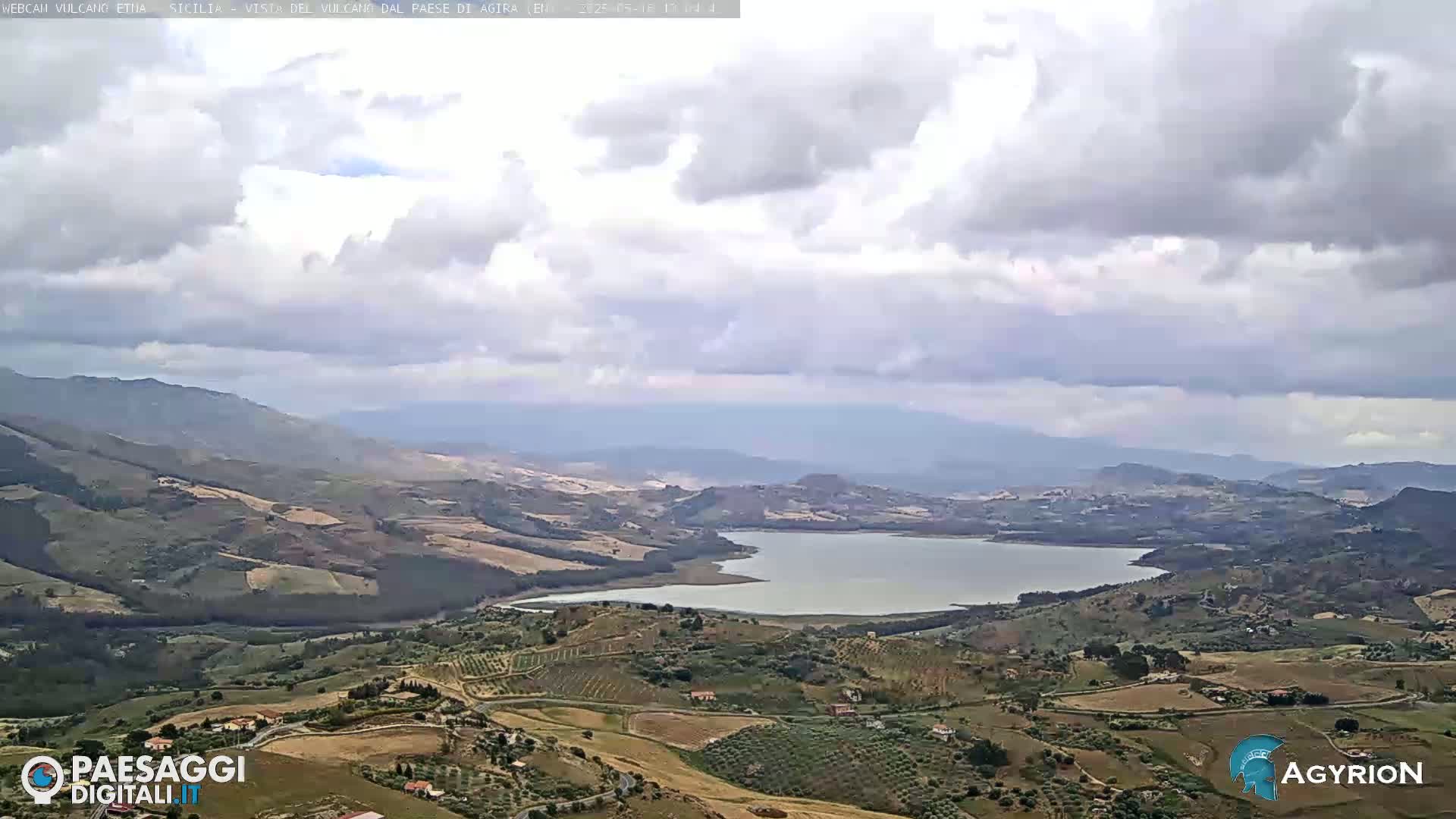 A partly cloudy day reveals a wide expanse of rolling hills and farmland surrounding a calm lake, with mountains visible in the distance.
