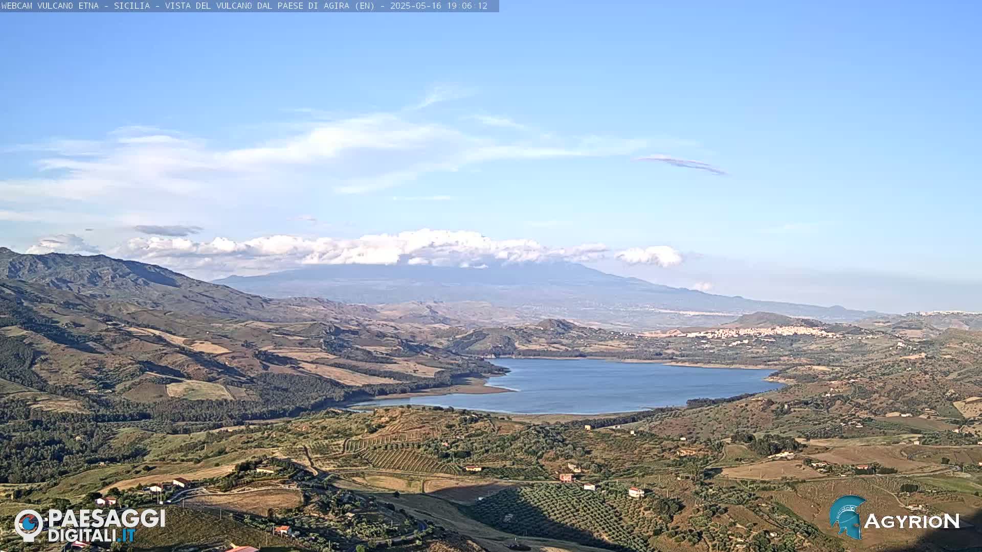A partly cloudy daytime view of a large lake nestled in a valley, with rolling hills and mountains in the background, one of which is a large volcano.