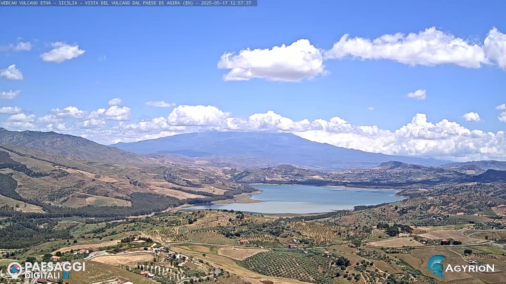 A partly cloudy, sunny day reveals a picturesque landscape of rolling hills, farmland, a lake, and a distant mountain range under a bright blue sky.