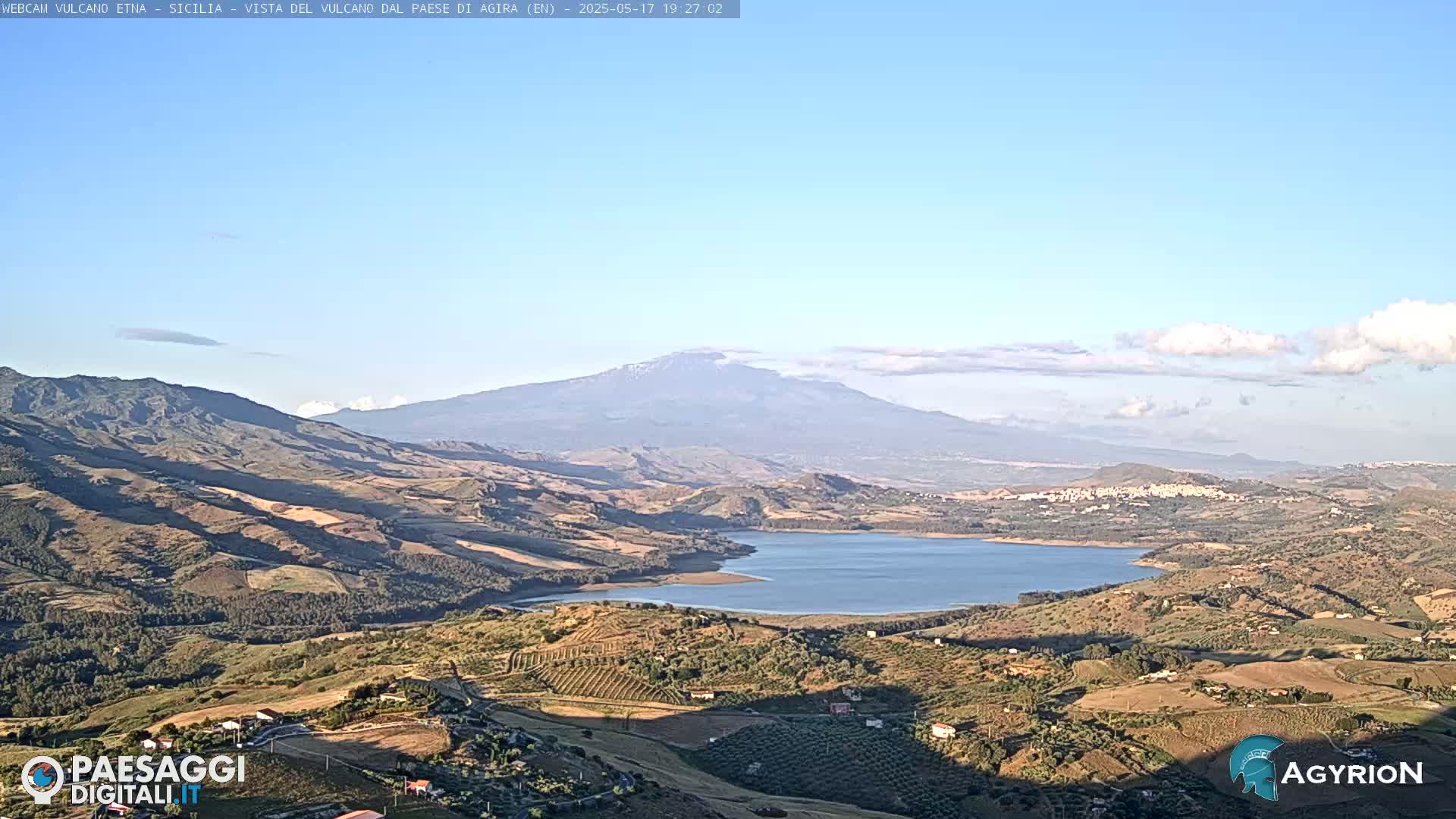 A distant, snow-capped mountain is visible over a valley with a lake, rolling hills, and farmland under a mostly clear sky.