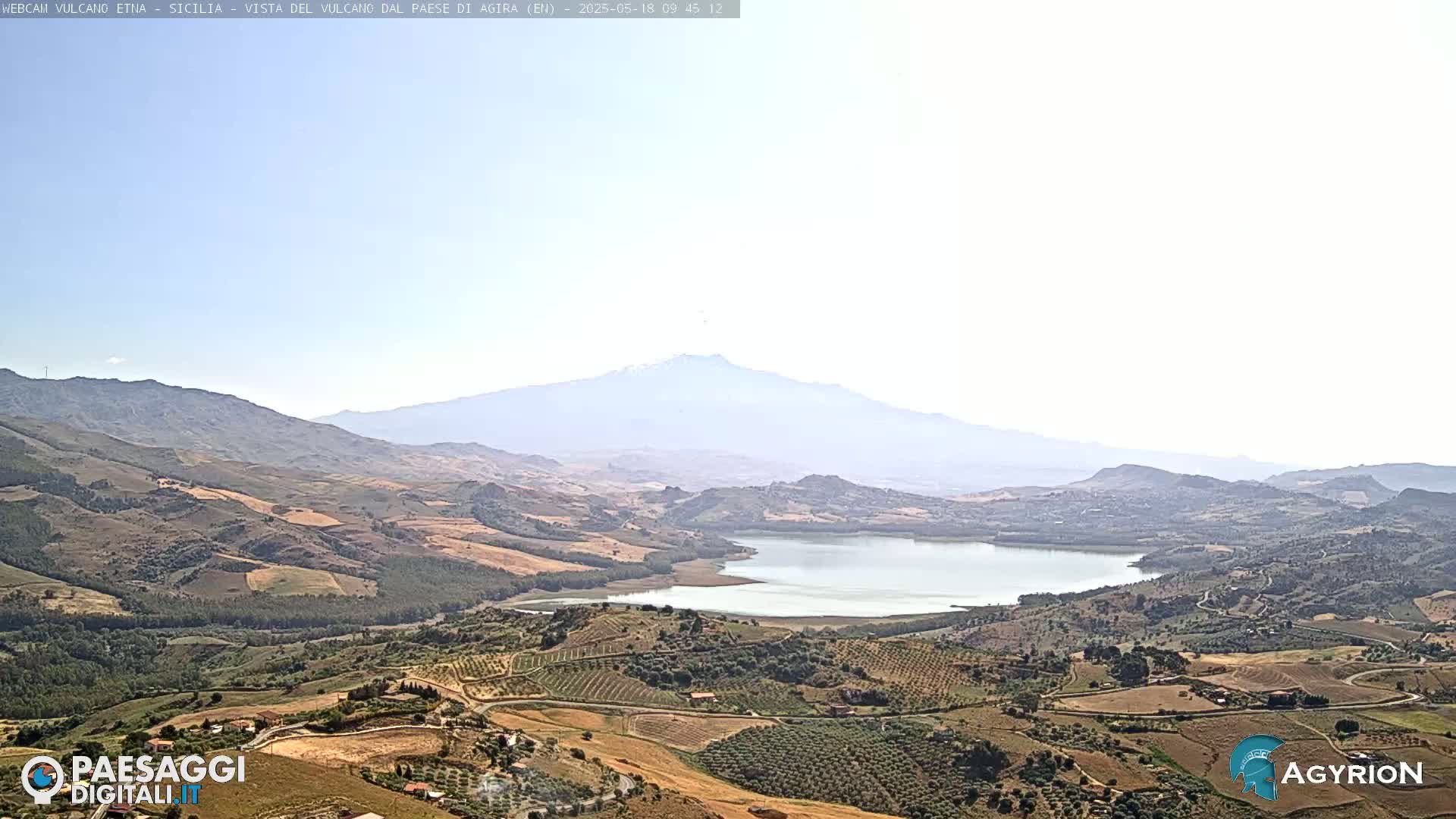 A hazy, sunny view of a valley with a lake and rolling hills, showcasing a distant, snow-capped mountain in the background.