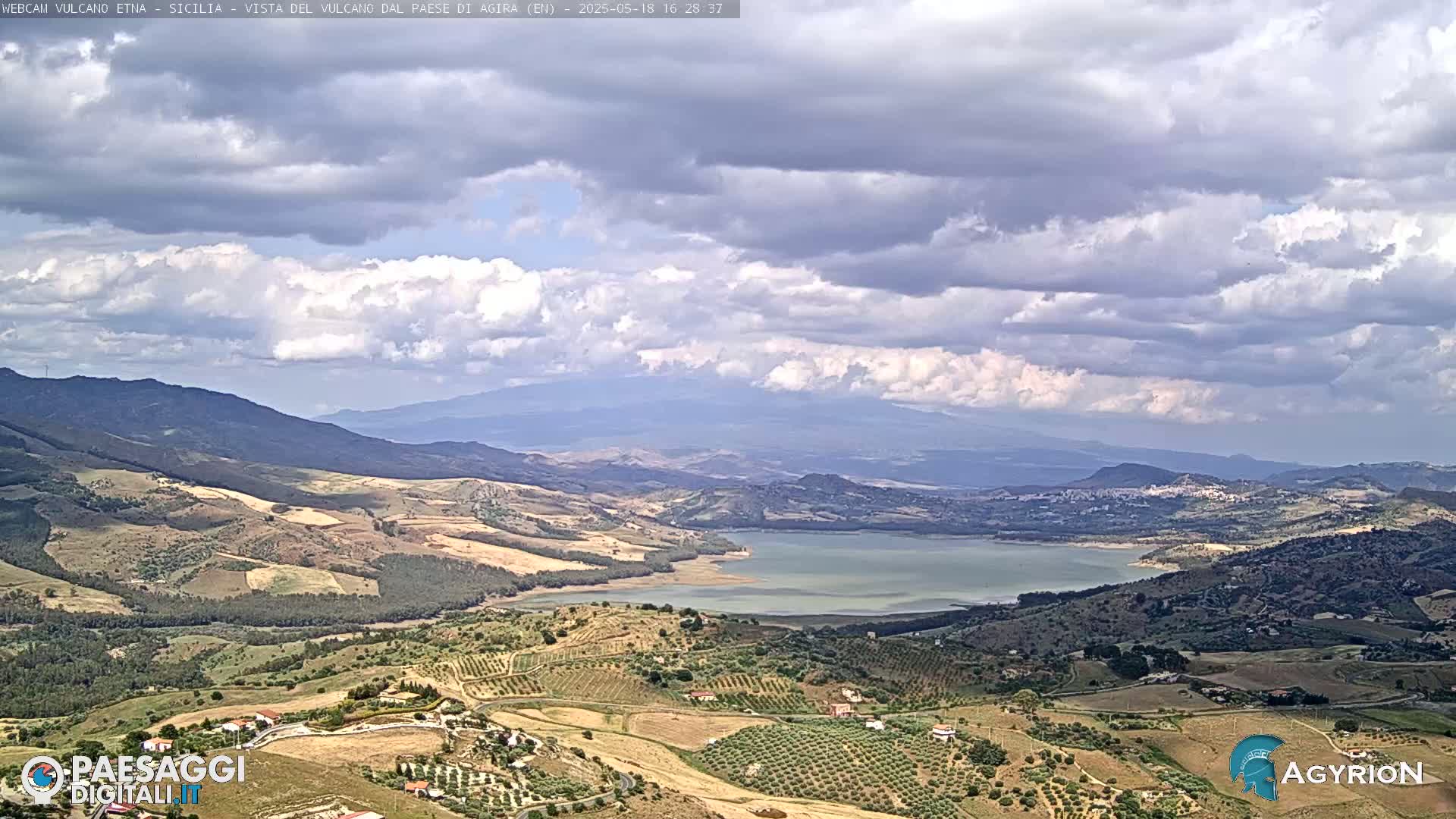 A partly cloudy day reveals a vast landscape of rolling hills, farmland, and a large lake nestled among mountains in the distance.