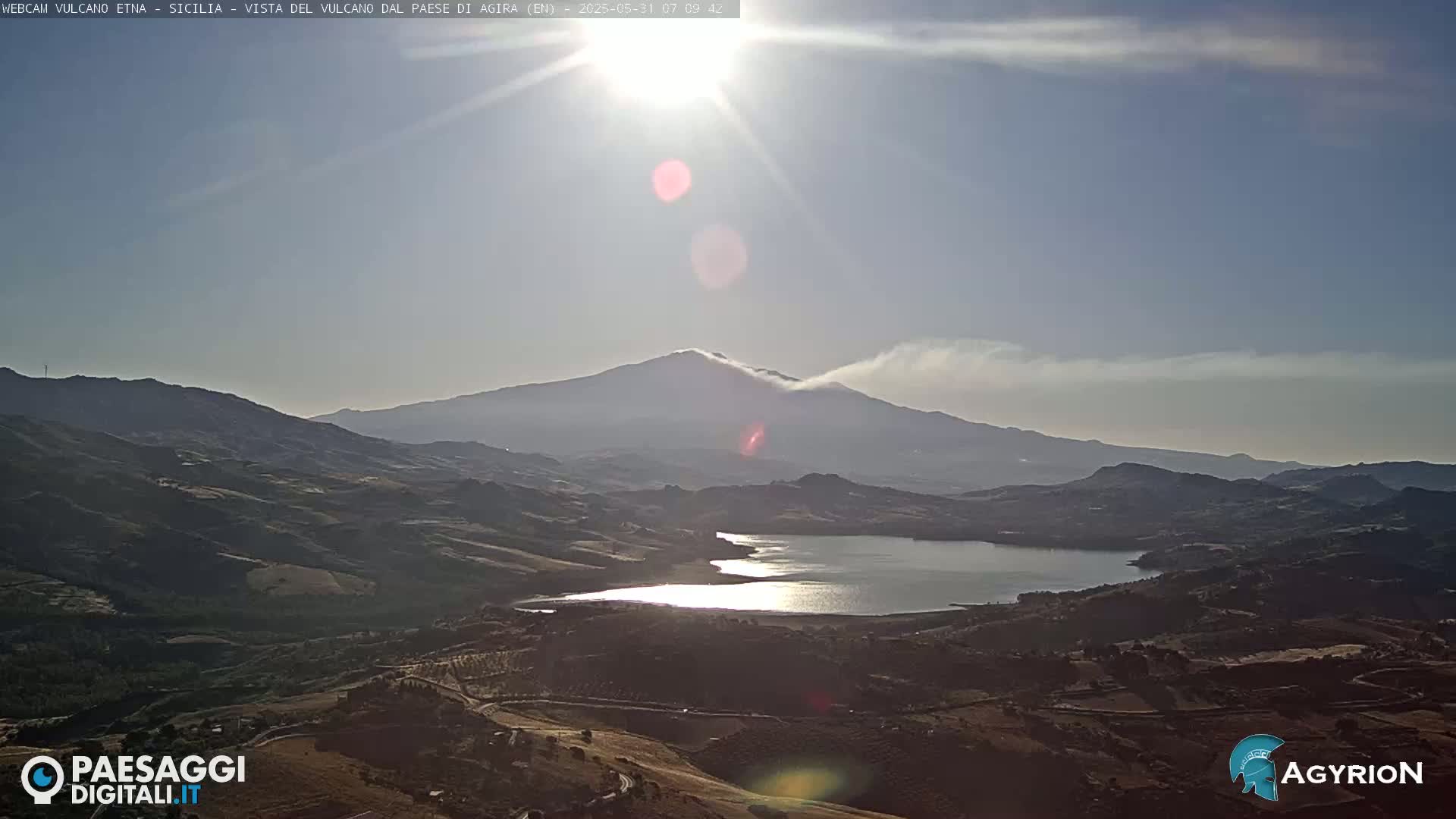 A sunlit view shows a large lake nestled in a valley below a distant, smoky volcano under a mostly clear sky.
