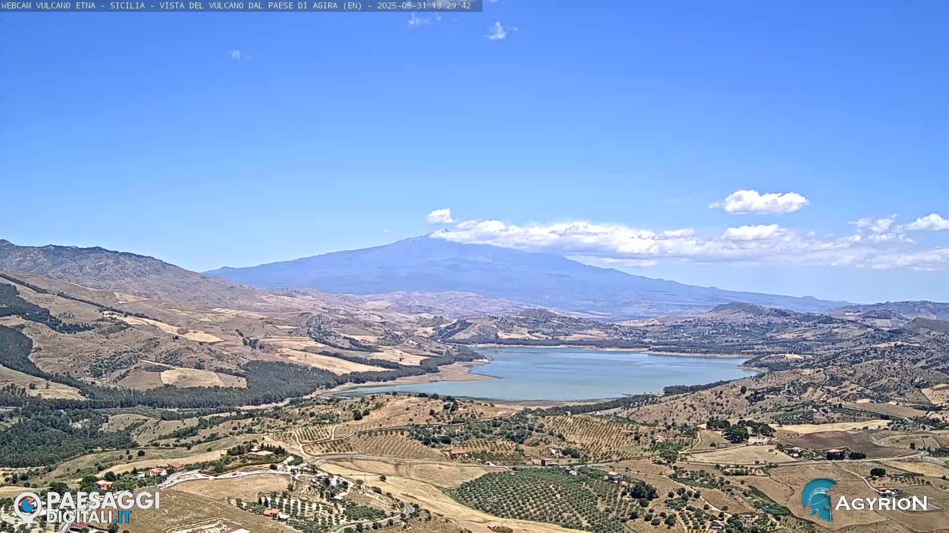 A mostly clear, sunny day reveals a large lake nestled in a valley, surrounded by arid, brown hills and olive groves, with Mount Etna in the background.