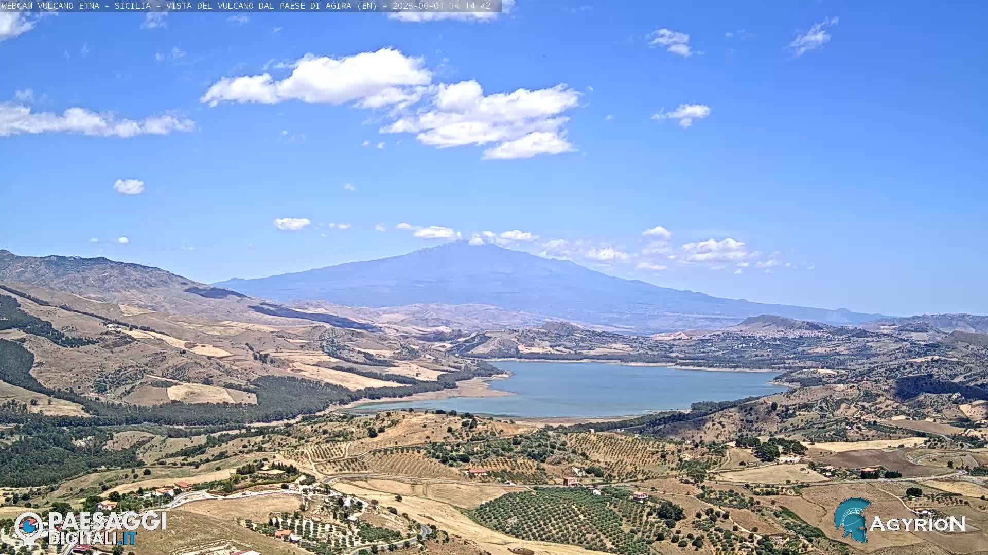 A large lake nestled in a valley below a distant, snow-capped mountain under a partly cloudy blue sky.