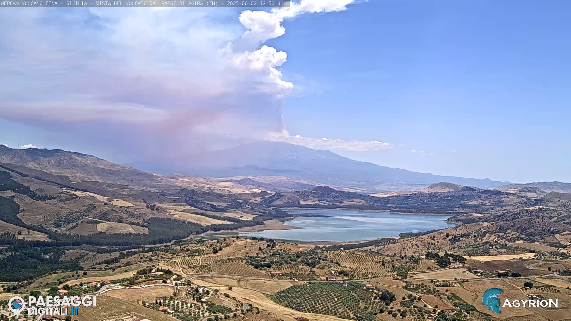 A large lake and expansive, dry, hilly landscape are seen under a mostly clear blue sky, with a volcano emitting a plume of smoke and ash in the distance.