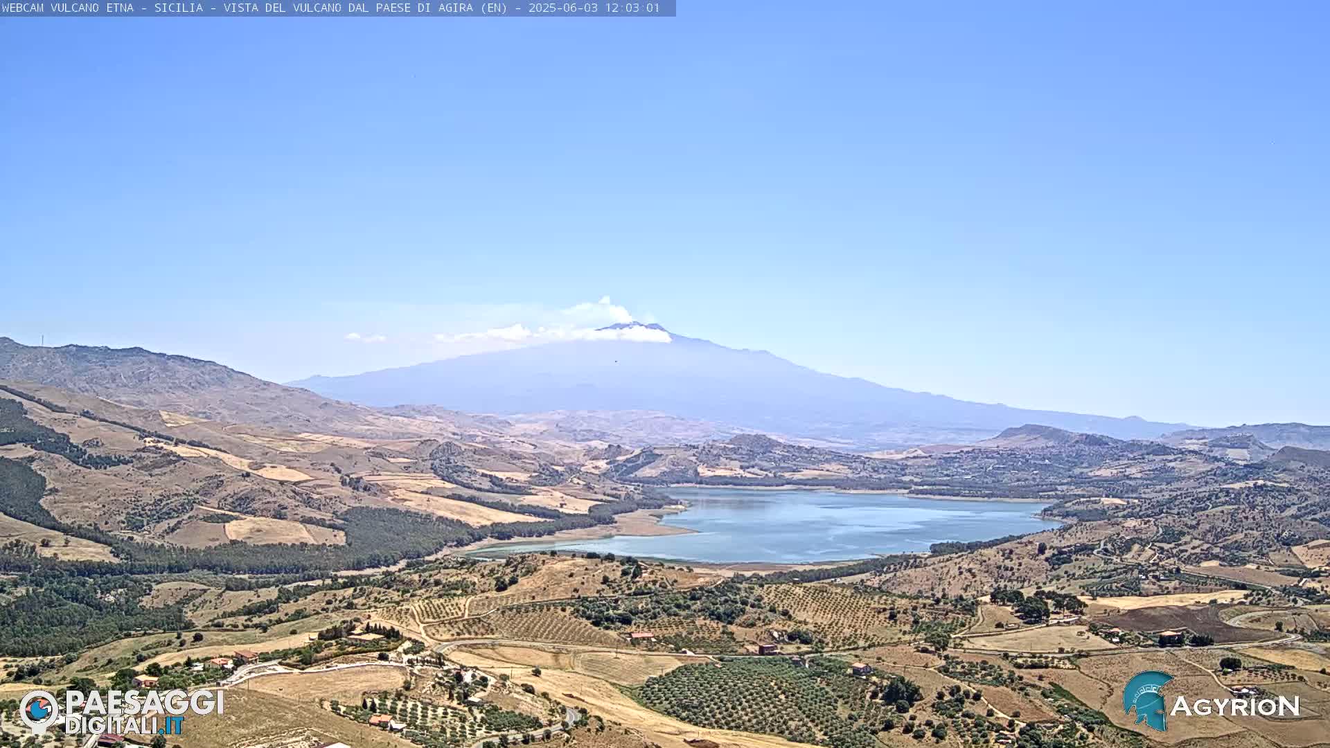 A vast, sunlit landscape features a large lake nestled among rolling hills, with Mount Etna and a small plume of smoke visible in the distance under a clear blue sky.