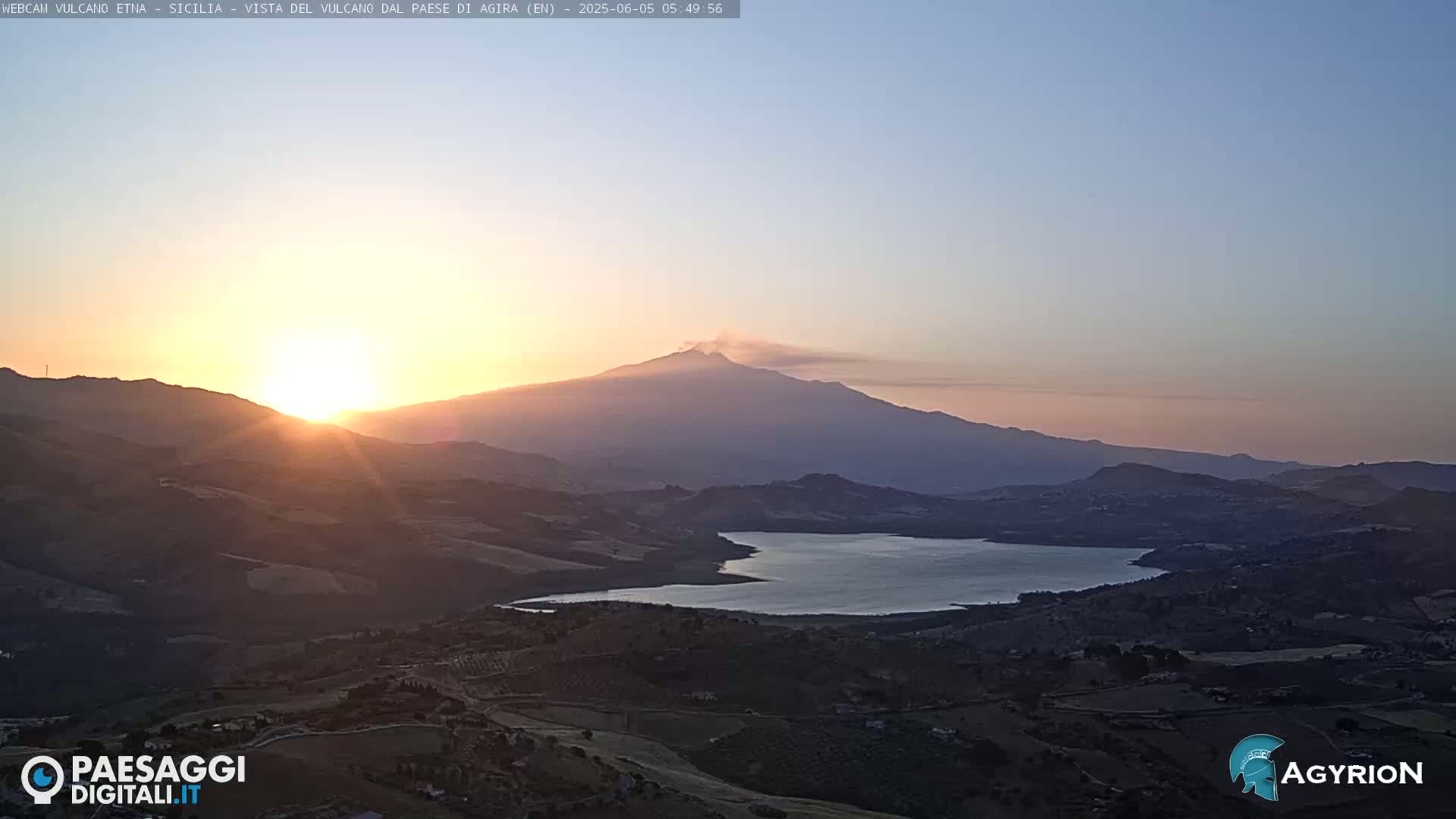 A sunlit vista reveals a lake nestled among rolling hills, with Mount Etna, emitting a plume of smoke, in the background at sunrise.